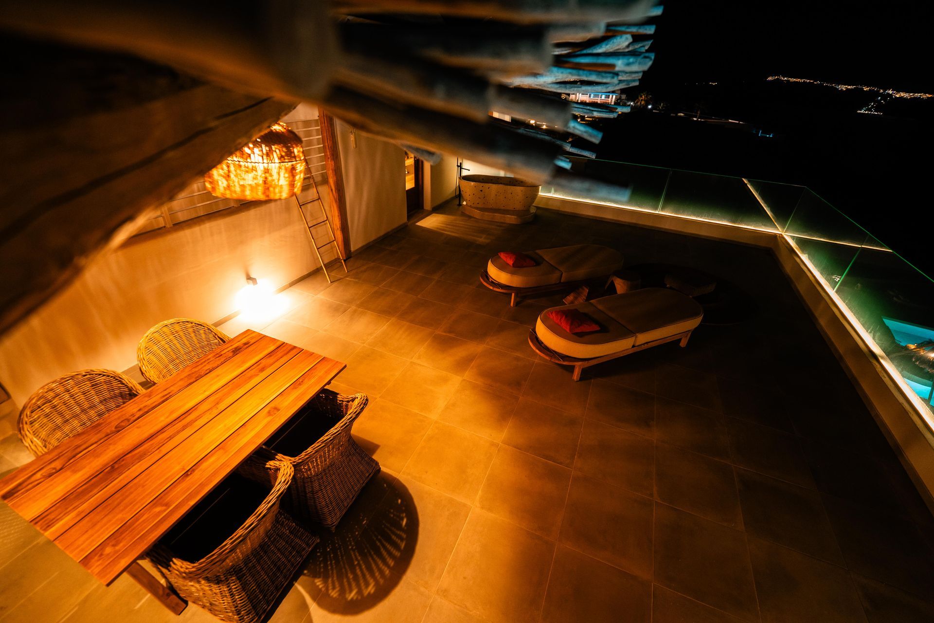Night view of a rooftop patio with table, chairs, and lounge beds overlooking a lit city.