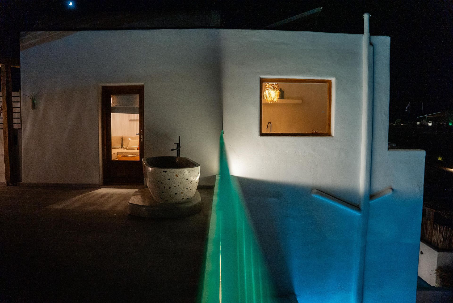 Exterior night shot of a white building with a lit window, door, and a bathtub on a patio.