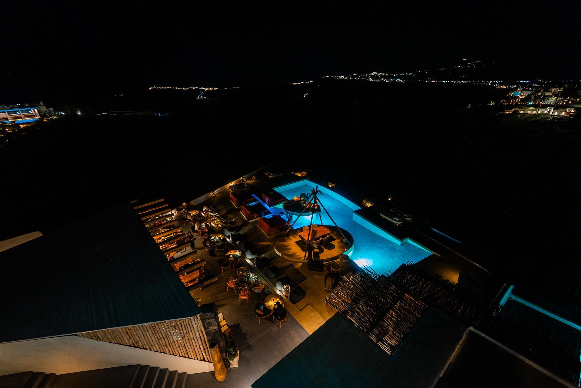 Nighttime aerial view of a pool and bar area, illuminated with blue and warm lights, city in the background.