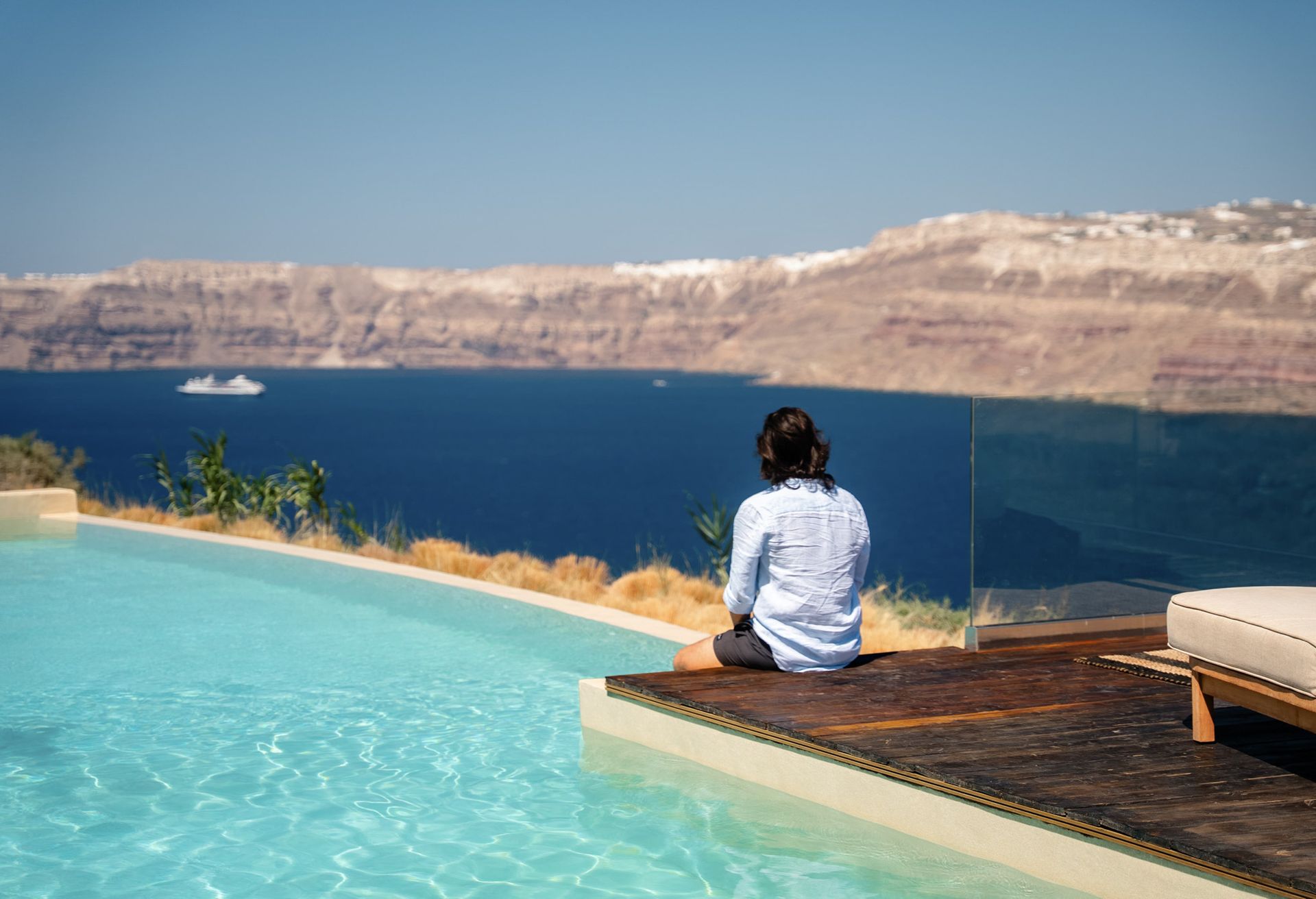 Person sitting by a pool, gazing at a blue sea and cliffs under a bright sky.
