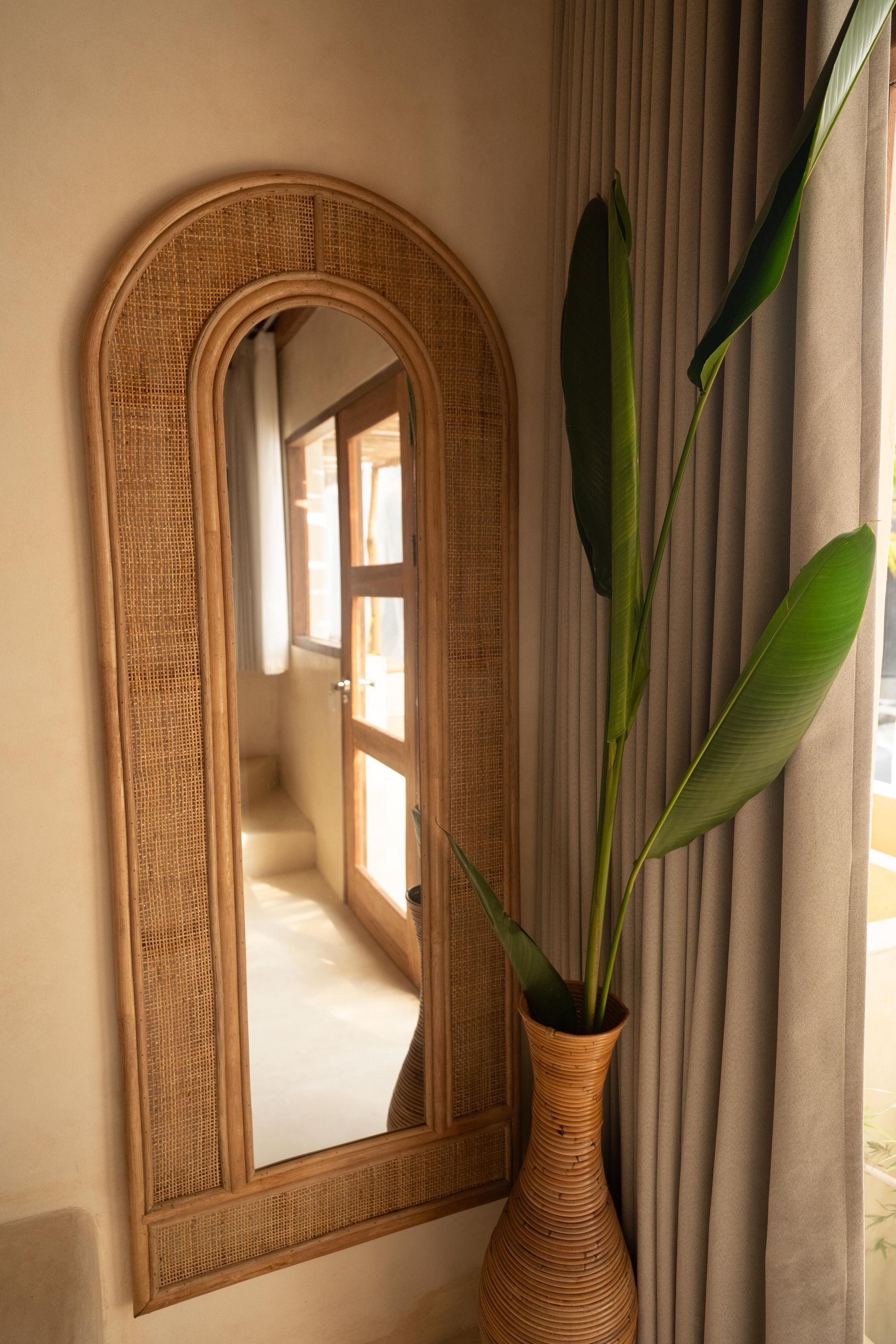 Woven-framed mirror reflecting a sunlit doorway, beside a tall vase with large green leaves and gray curtains.