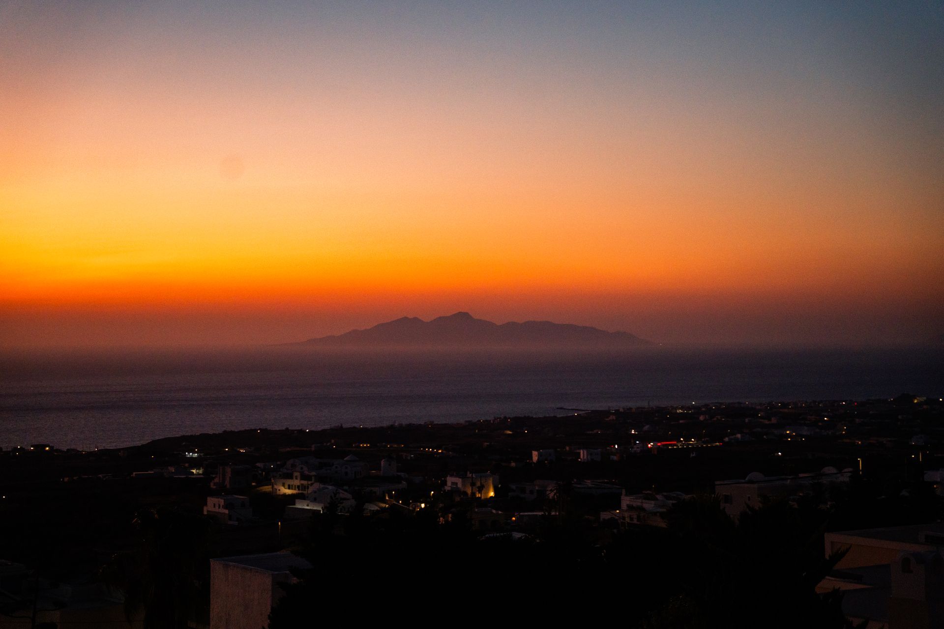 Sunset over a coastal city, with orange sky and a dark mountain silhouette on the horizon