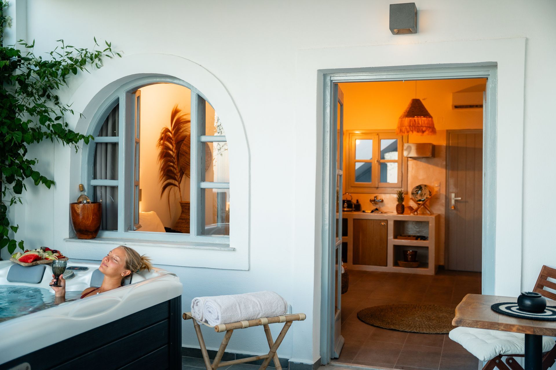 Woman in a jacuzzi, looking at a room with a kitchenette. White walls, blue sky, warm interior lighting.