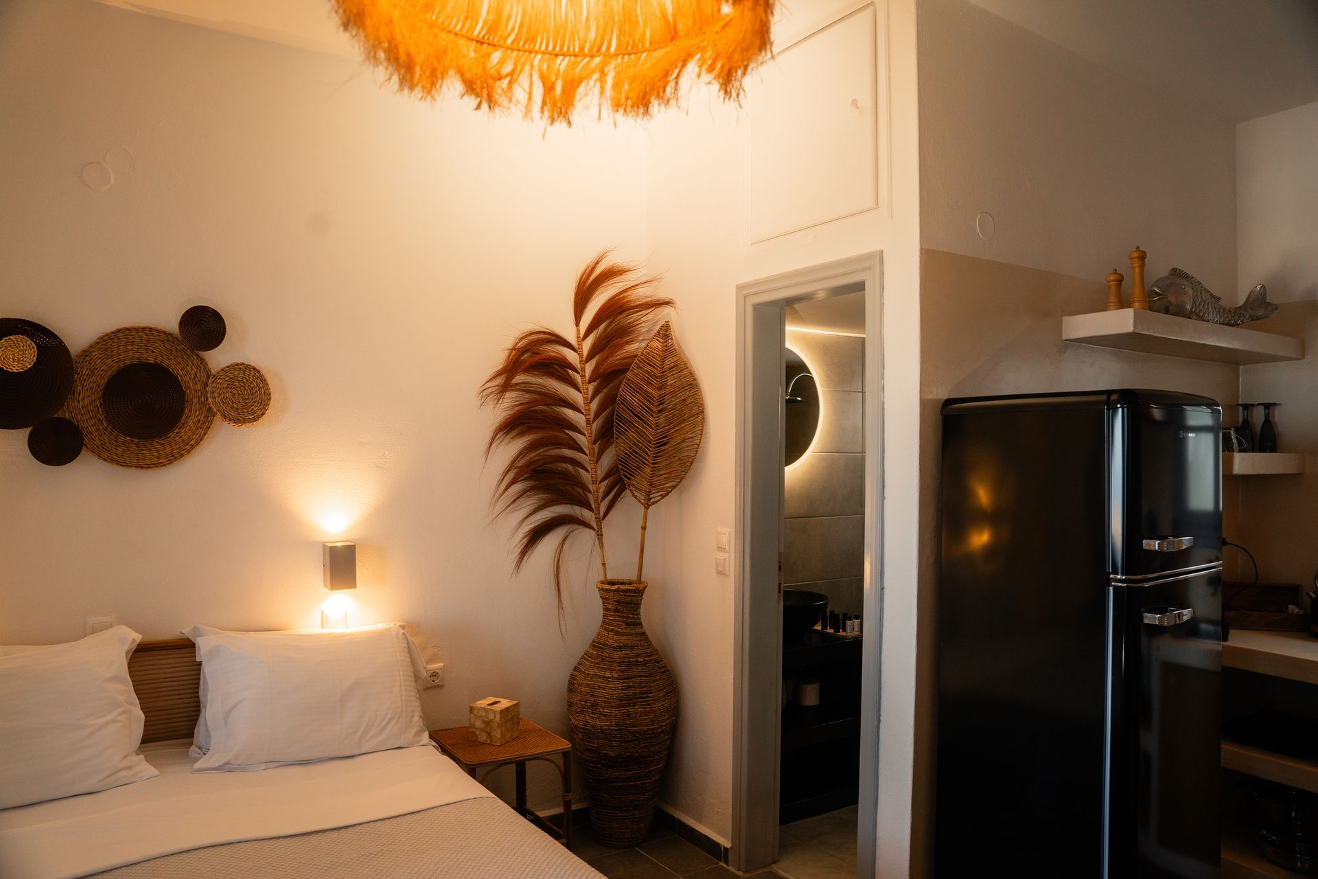 Cozy bedroom with double bed, tall vase with plumes, black refrigerator, and bathroom visible.