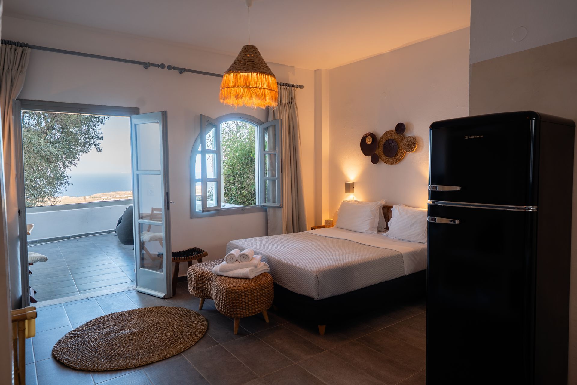 Bedroom with bed, black fridge, open door to a balcony with sea view, neutral colors.