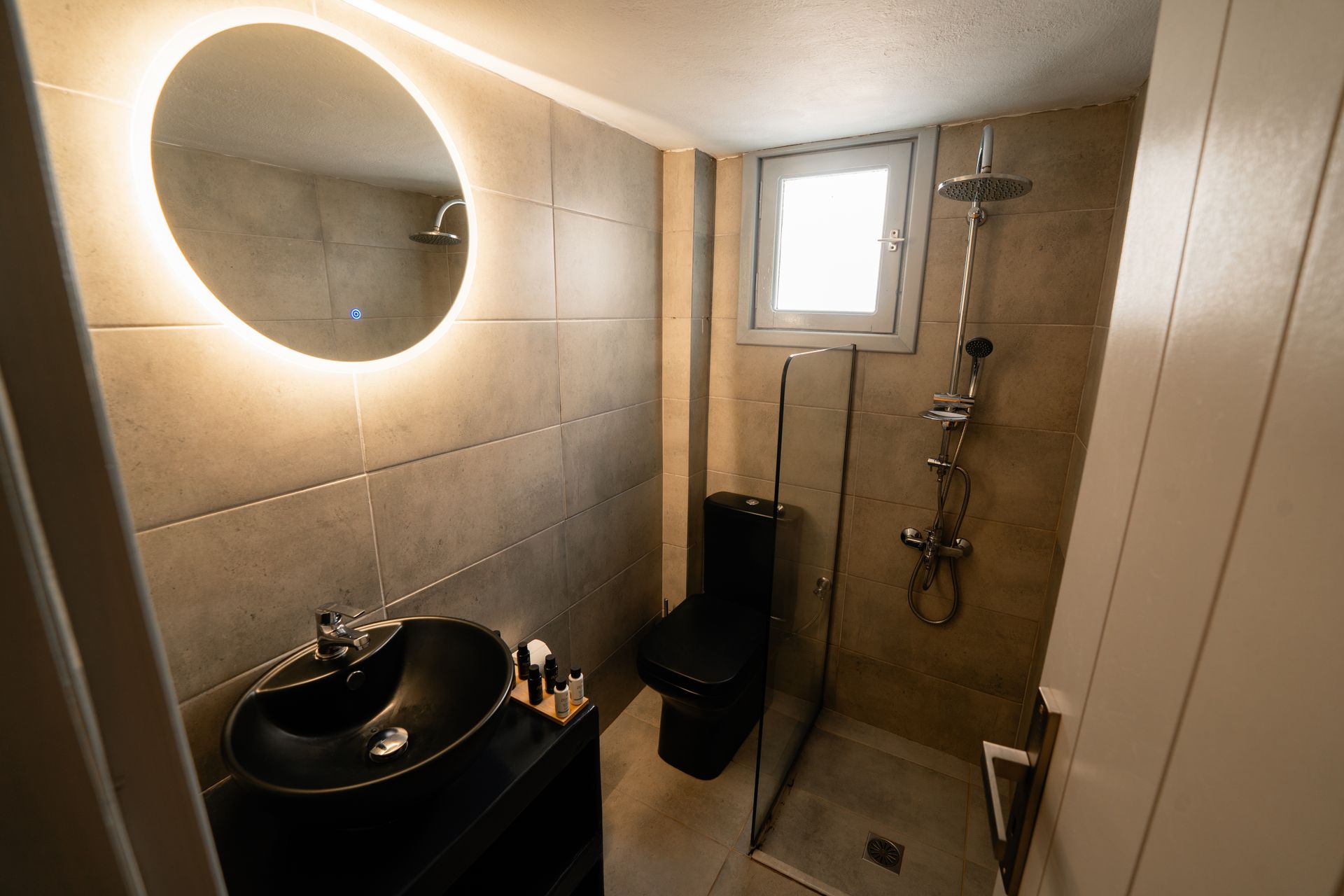 Small bathroom with a black sink, toilet, and shower, and a backlit circular mirror.