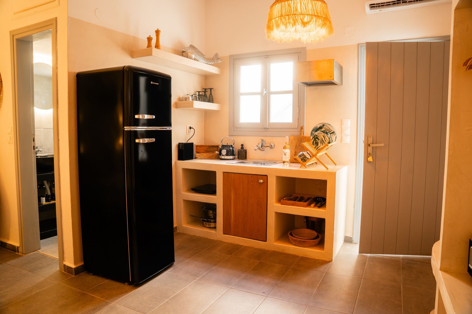 Black retro refrigerator in a sunlit kitchen with a built-in counter, shelves, and a small window.