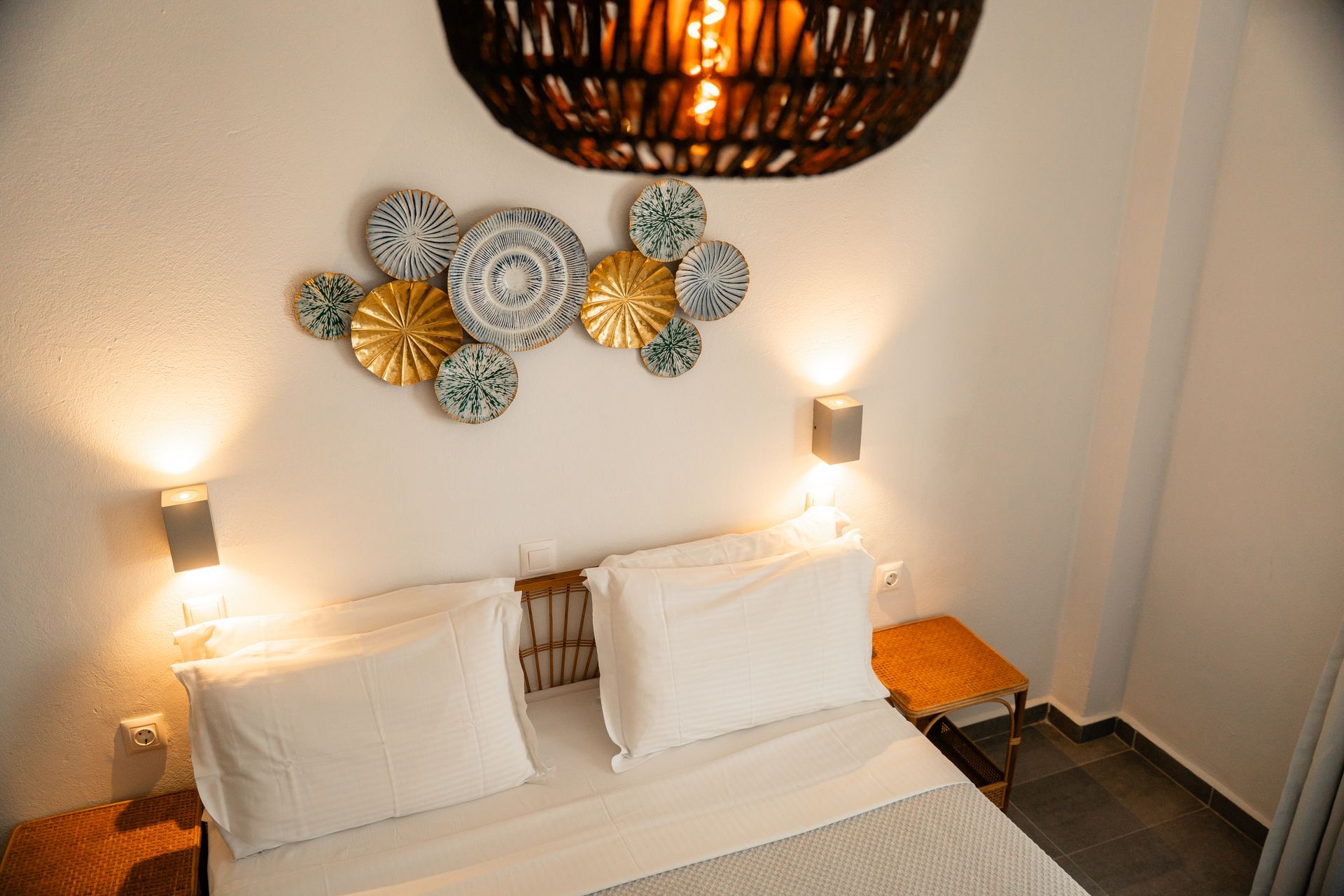 Bedroom with bed, decorative plates on the wall, bedside tables, wall sconces, and a woven lampshade.