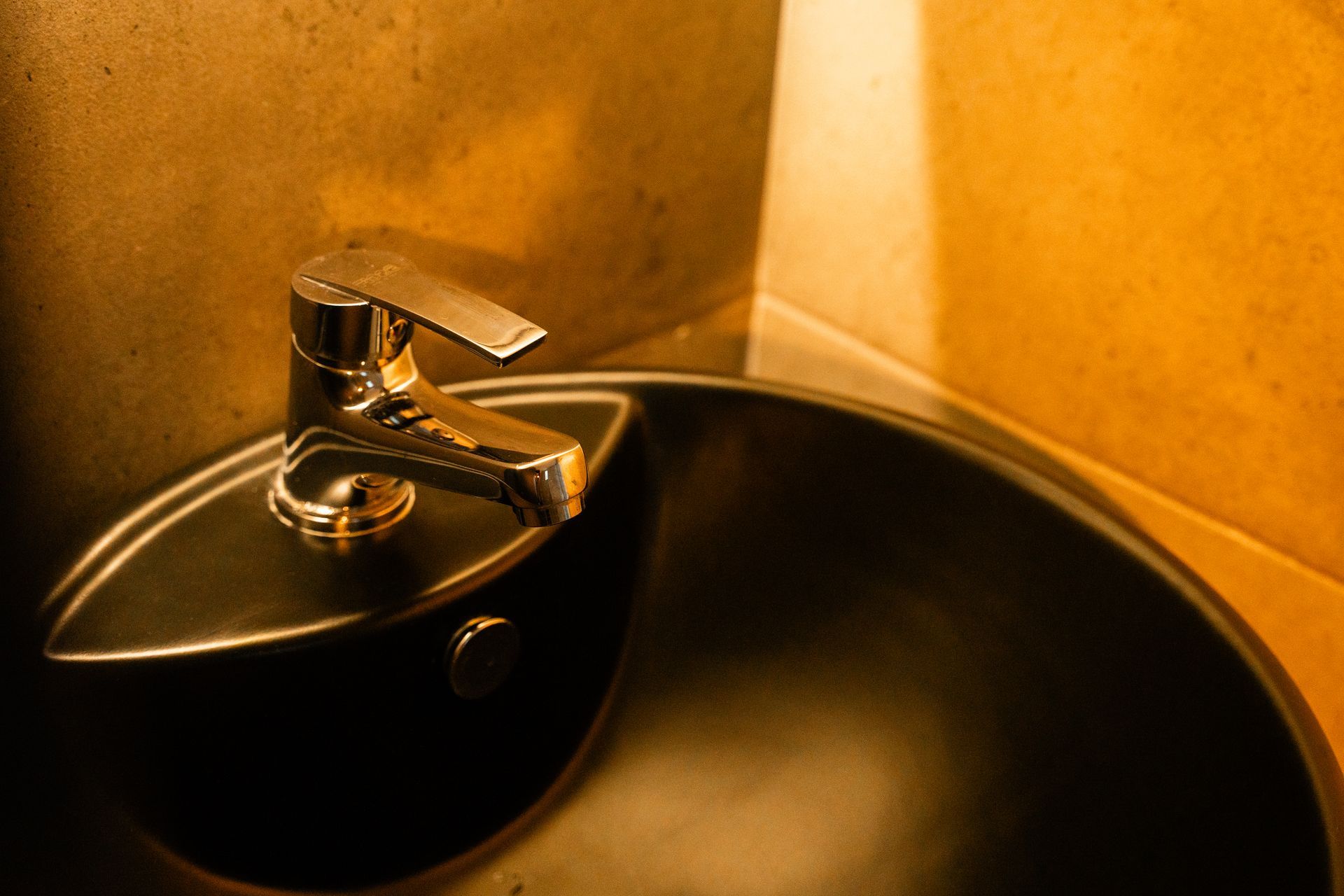 Faucet over a dark, rounded sink in a corner, lit with warm tones.