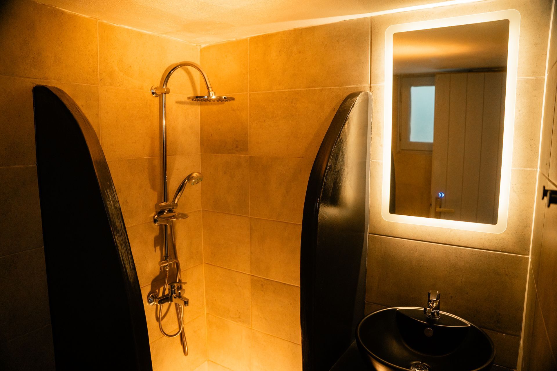 Shower area with gold-toned tiles, a curved divider, silver fixtures, and a backlit mirror above a black sink.