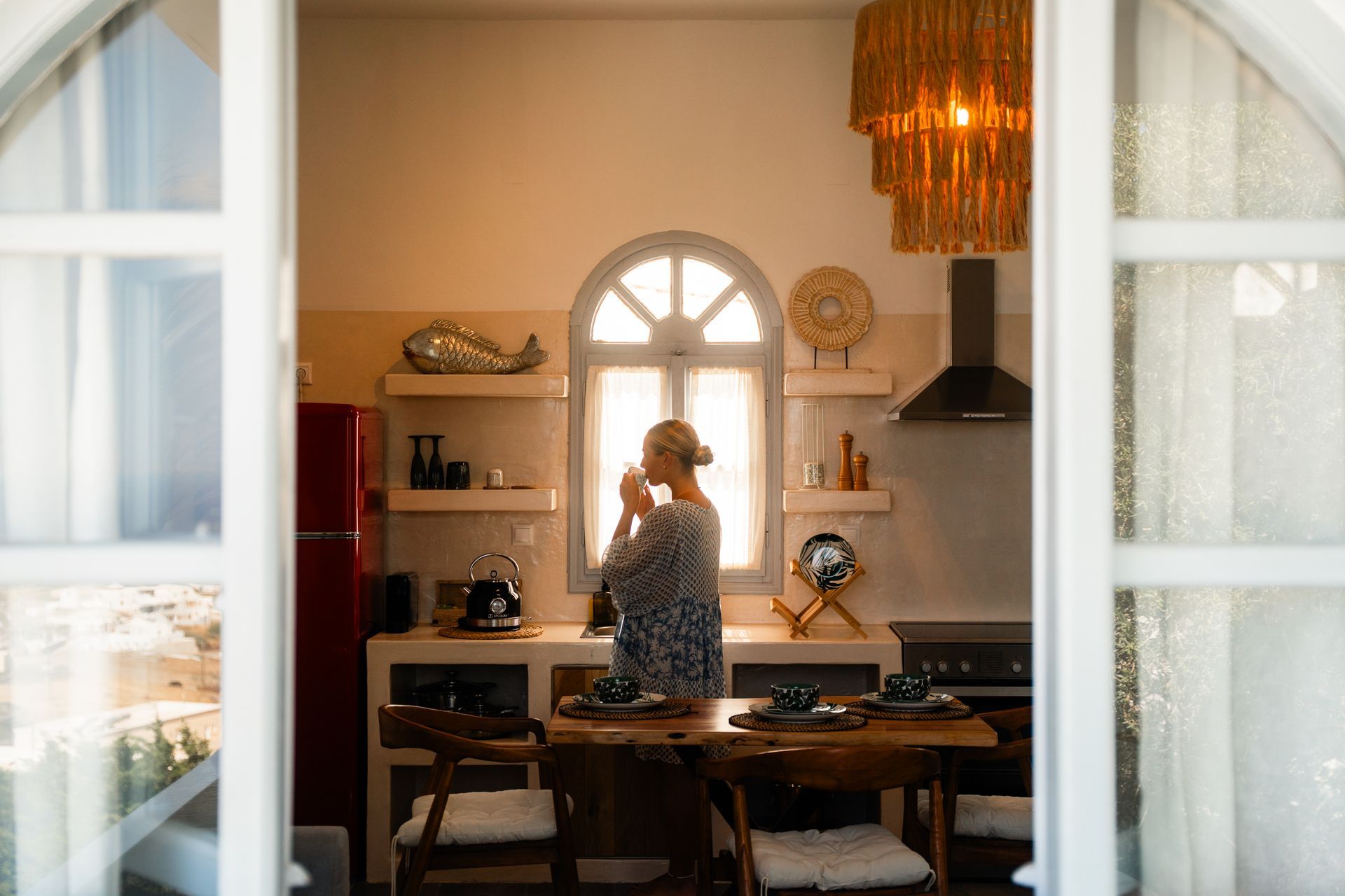 Woman sipping coffee in a sunlit kitchen, seen through open white doors.
