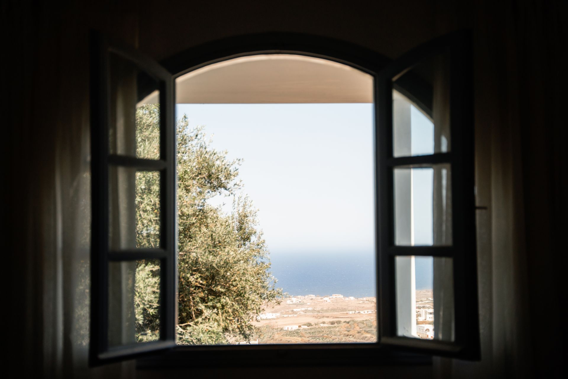 Open window frames a view of a tree, the sea, and a distant landscape on a sunny day.
