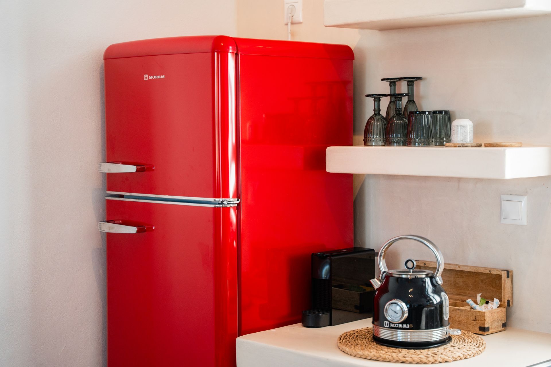 Red retro refrigerator in a white kitchen, with a kettle, coffee machine, and shelf with glasses.