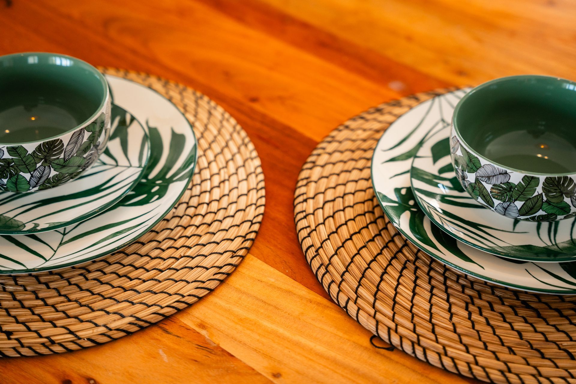 Two place settings with green bowls, plates with leaf design, on round woven placemats, on a wooden table.