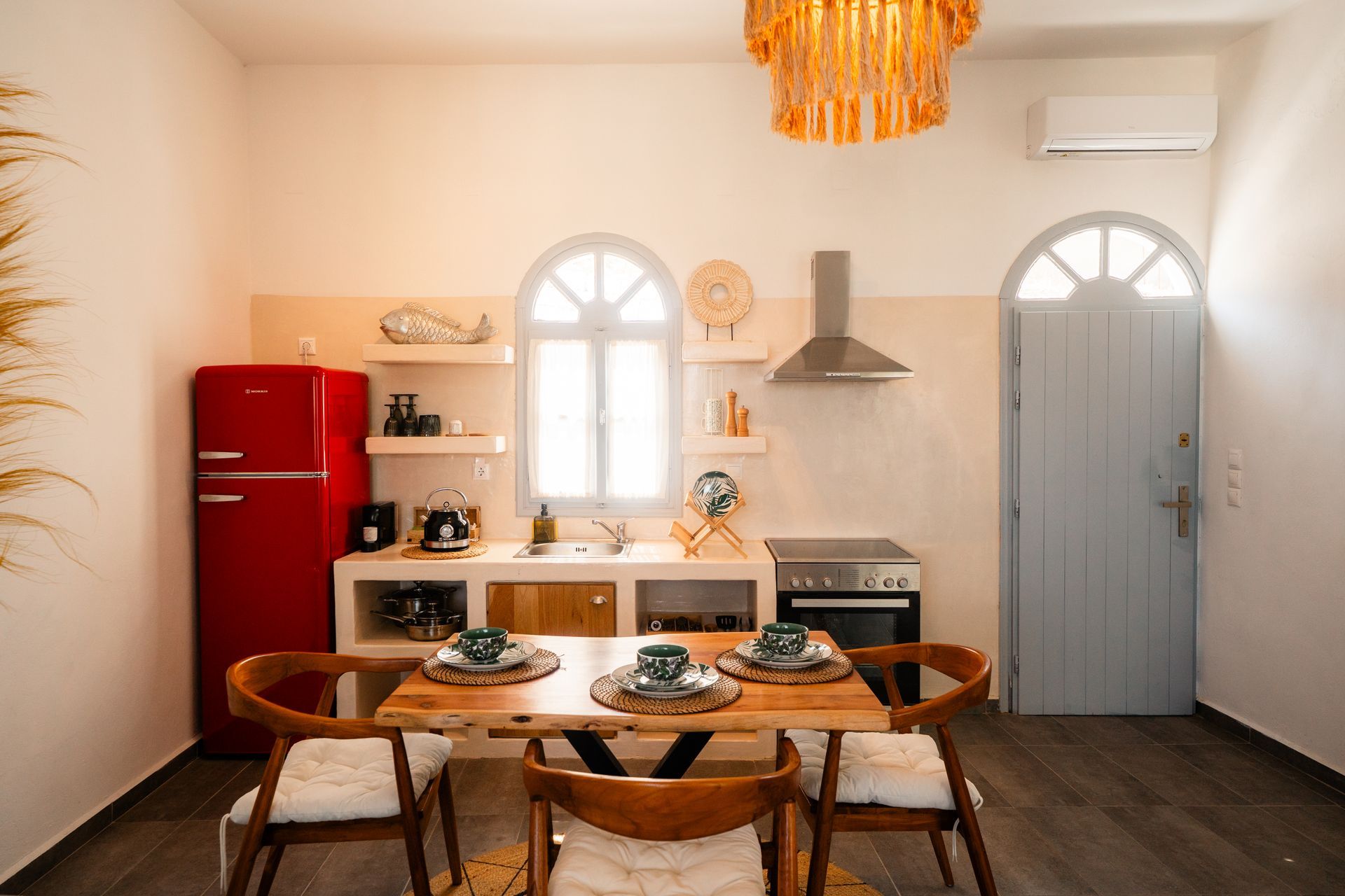 A well-lit kitchen with a red fridge, wooden table, and arched window. Neutral tones with bright accents.