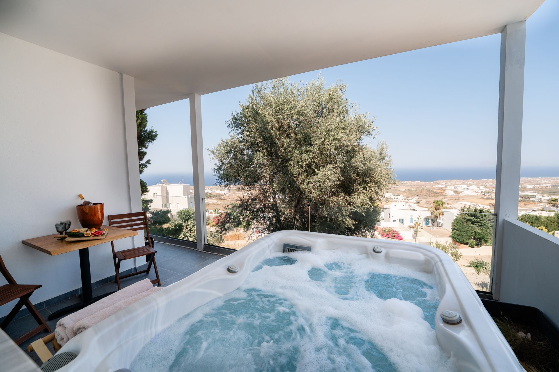 A balcony with a hot tub overlooking a cityscape and ocean. Table with chairs and tree in view.