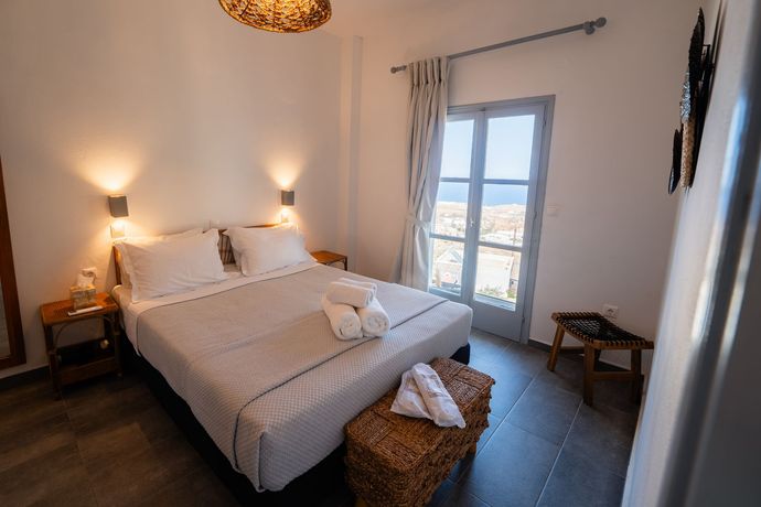 Cozy bedroom with a made bed, warm lamps, wicker bench, and balcony doors opening to a bright view