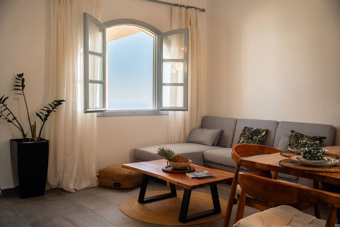 Bright living room with gray sofa, wood furniture, open window with sea view.