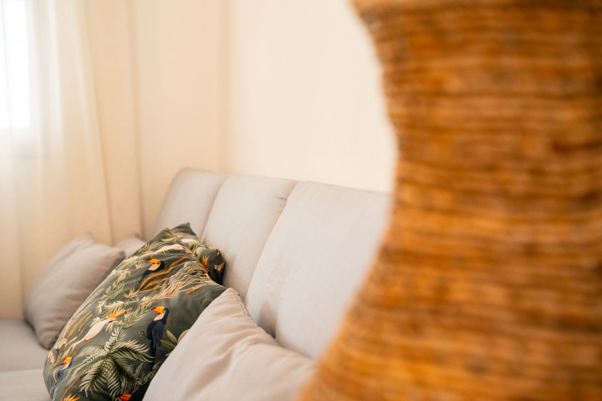 Beige sofa with pillows, patterned with toucans, near a textured brown vase.