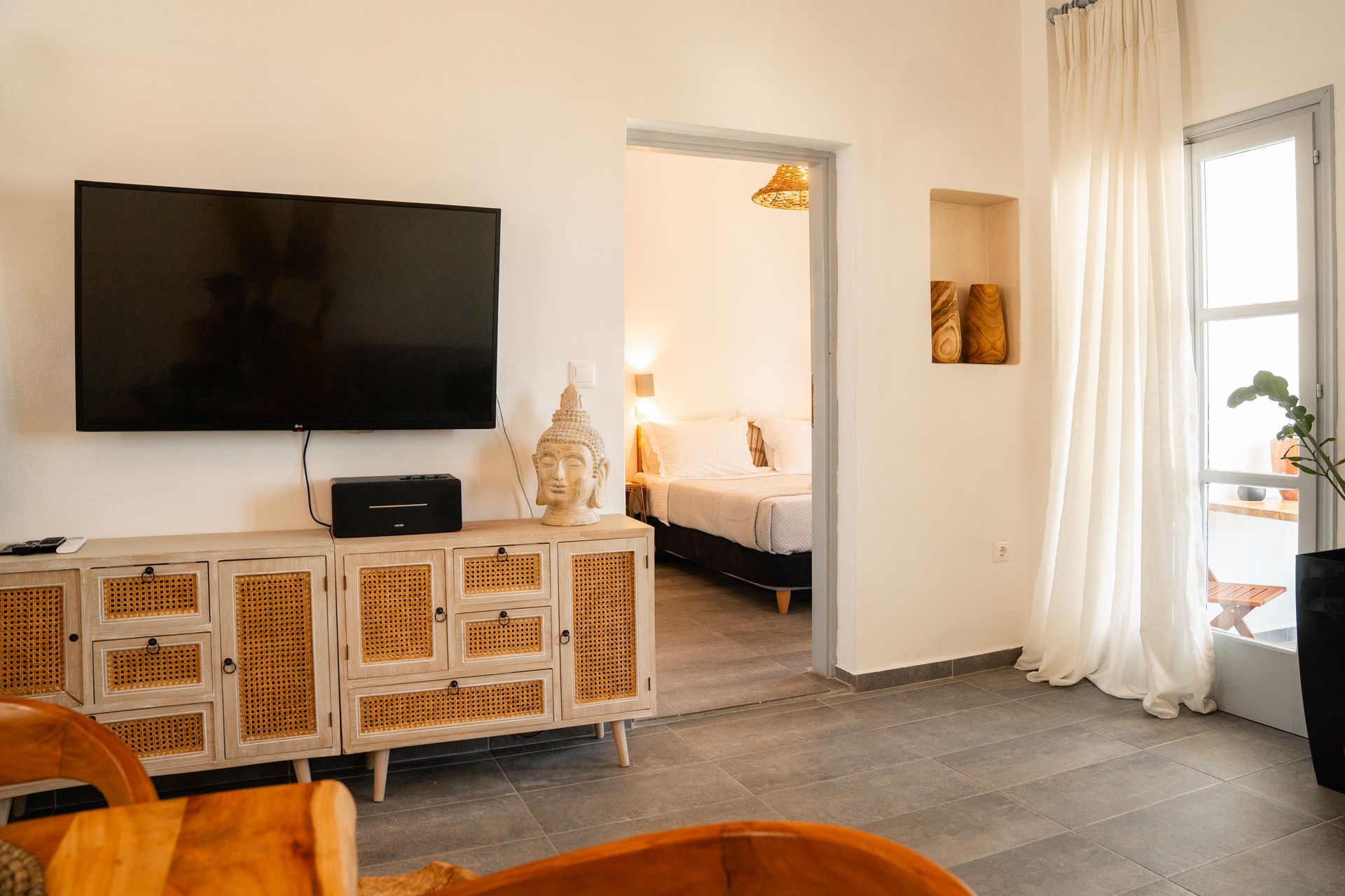 Living room with TV, wooden cabinet, and open doorway to a bedroom, with a window and white curtains.