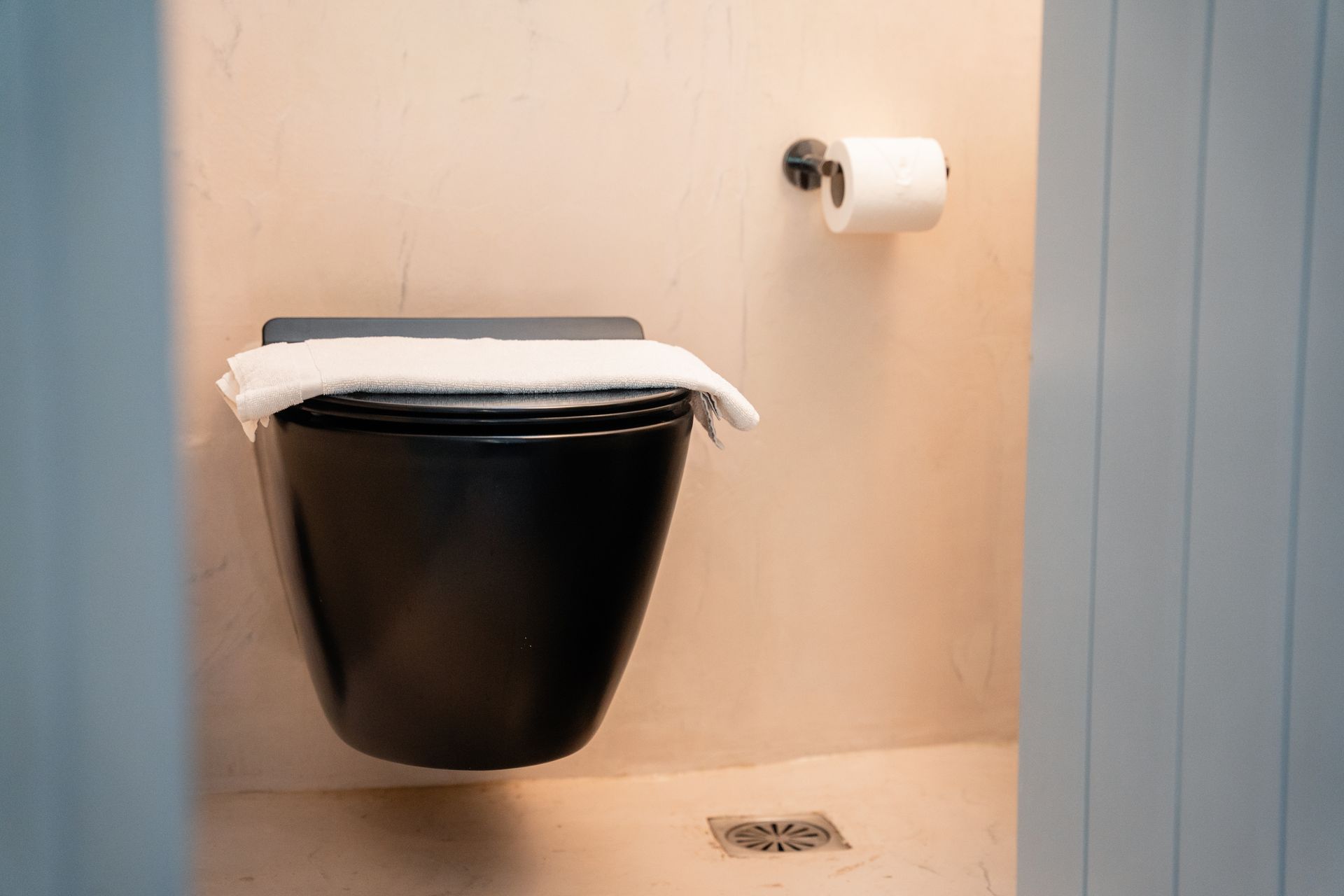 Black toilet with a towel on the seat, roll of toilet paper on the wall, and a floor drain.