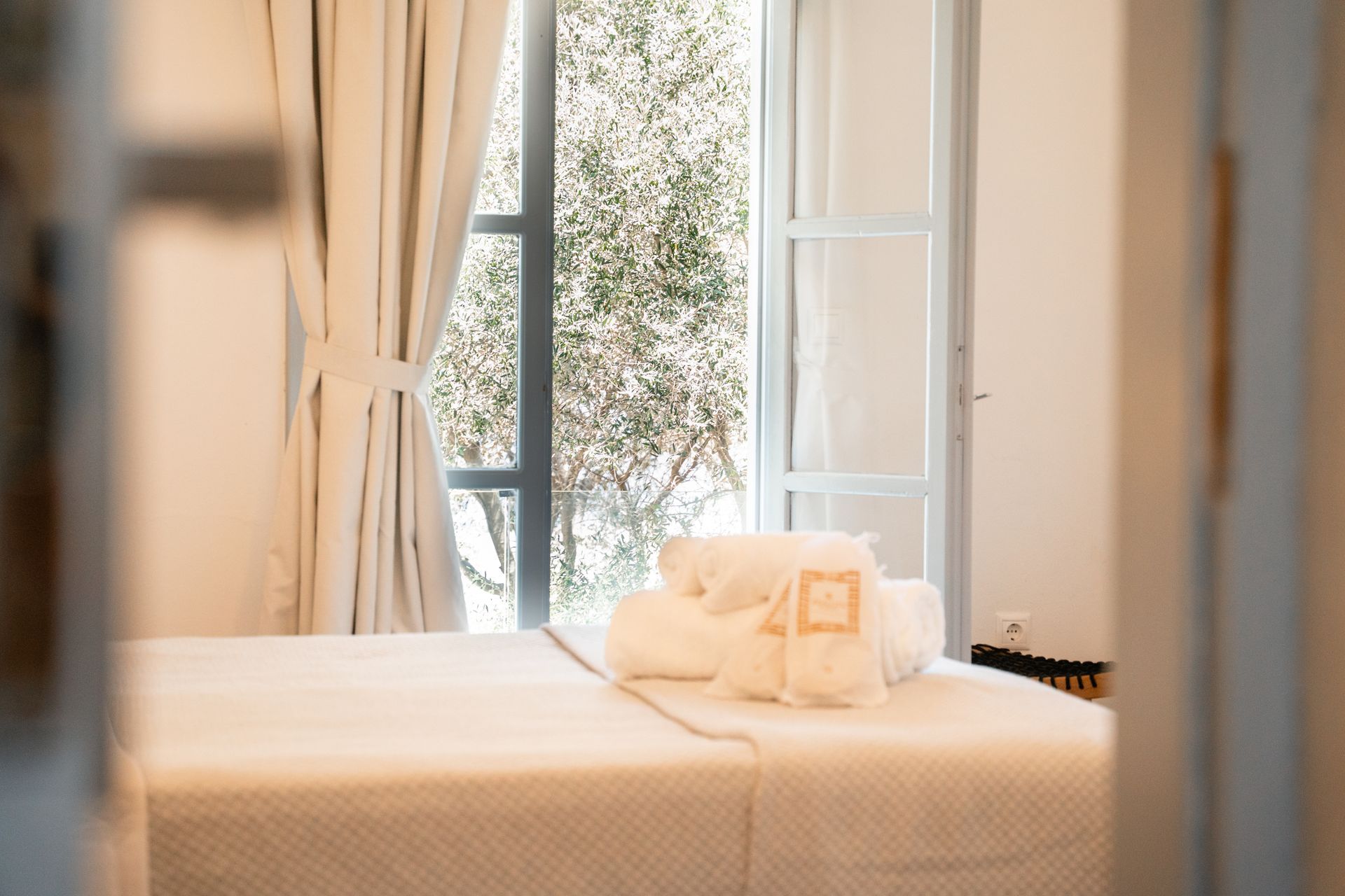 Massage table with white linens near an open window, sunlight, towels, and curtains.