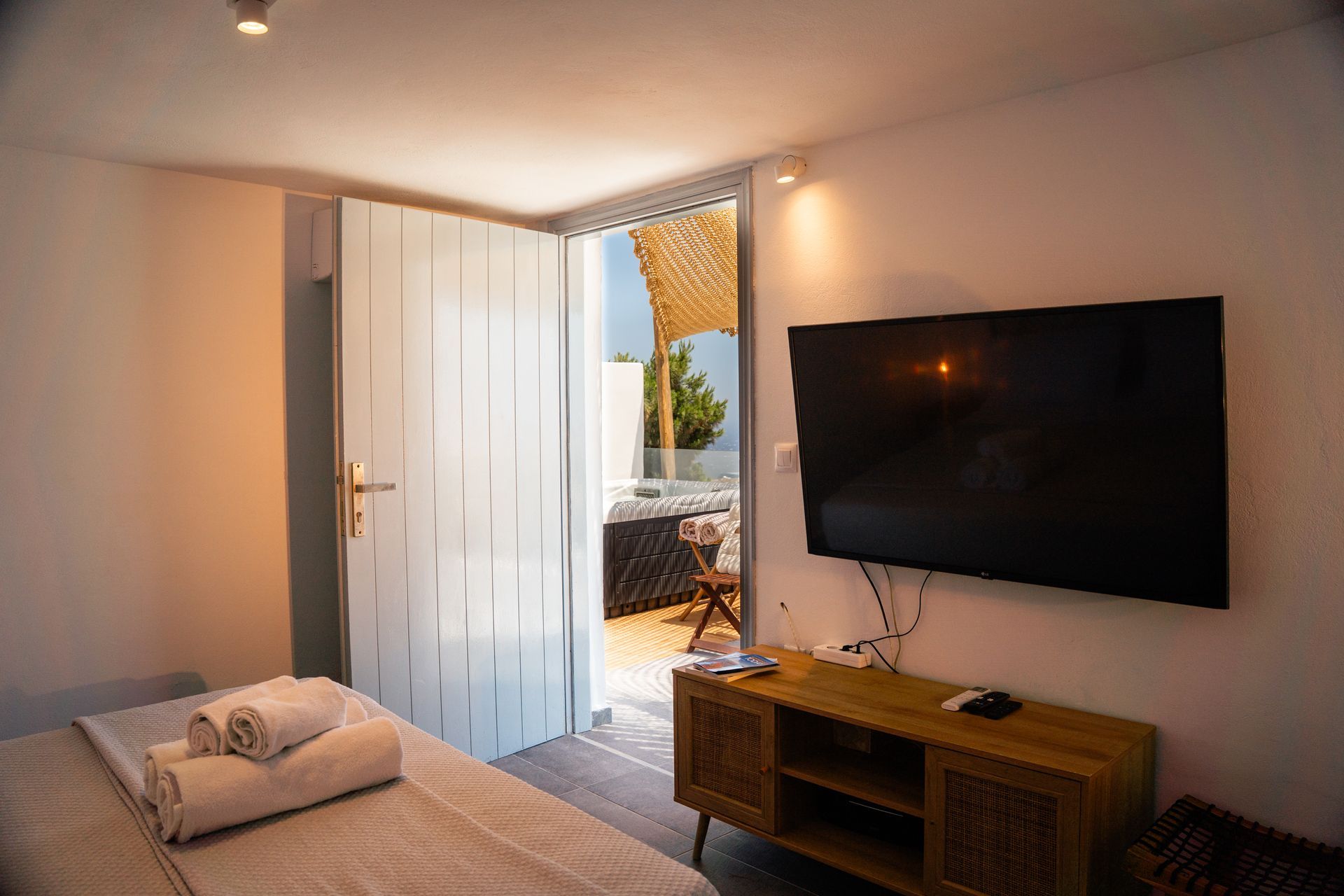 Bedroom with a large TV, open door to patio with a table and chairs, and a bed with towels.