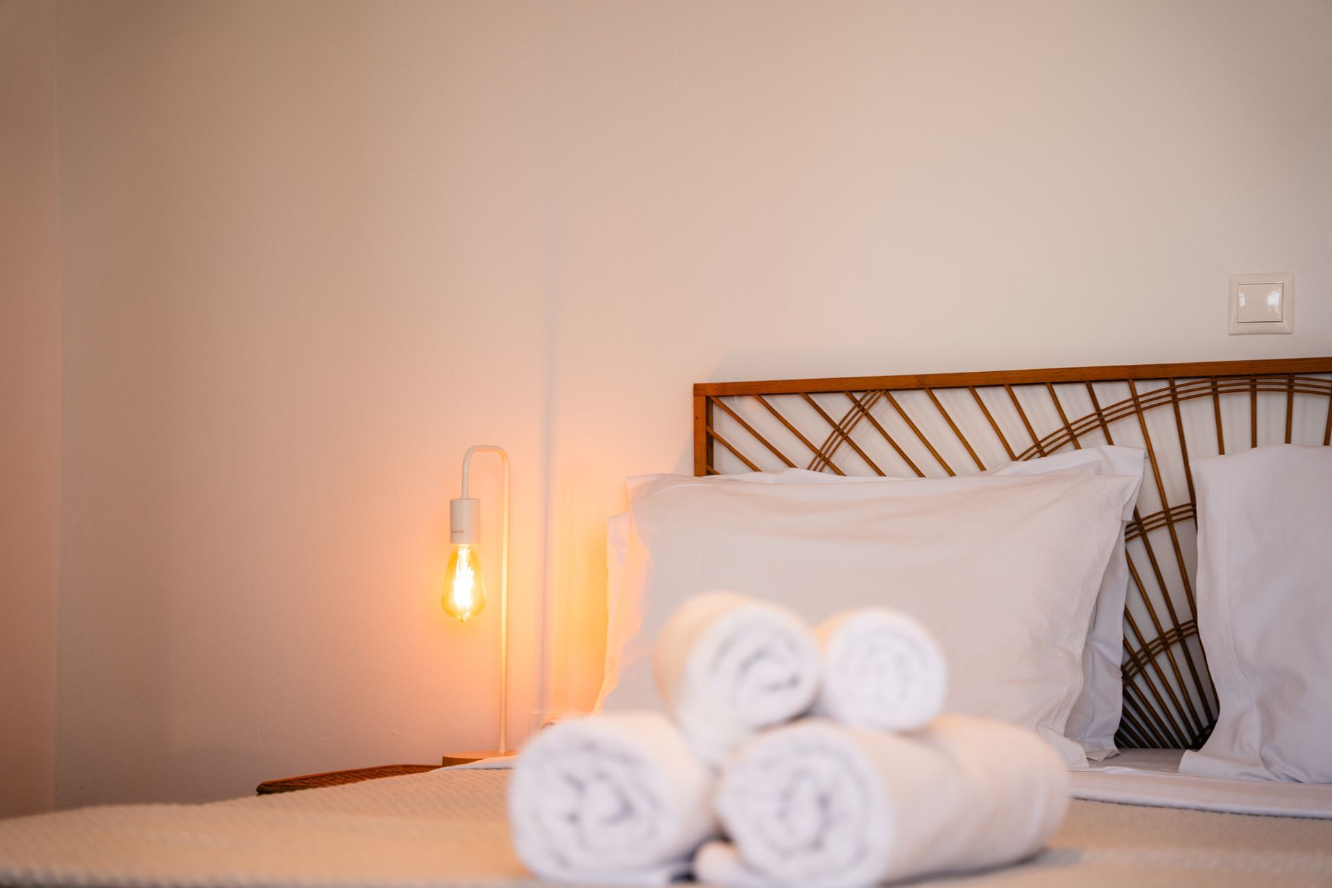 Bedroom with white linens, rolled towels, bamboo headboard, and a warm-toned light.