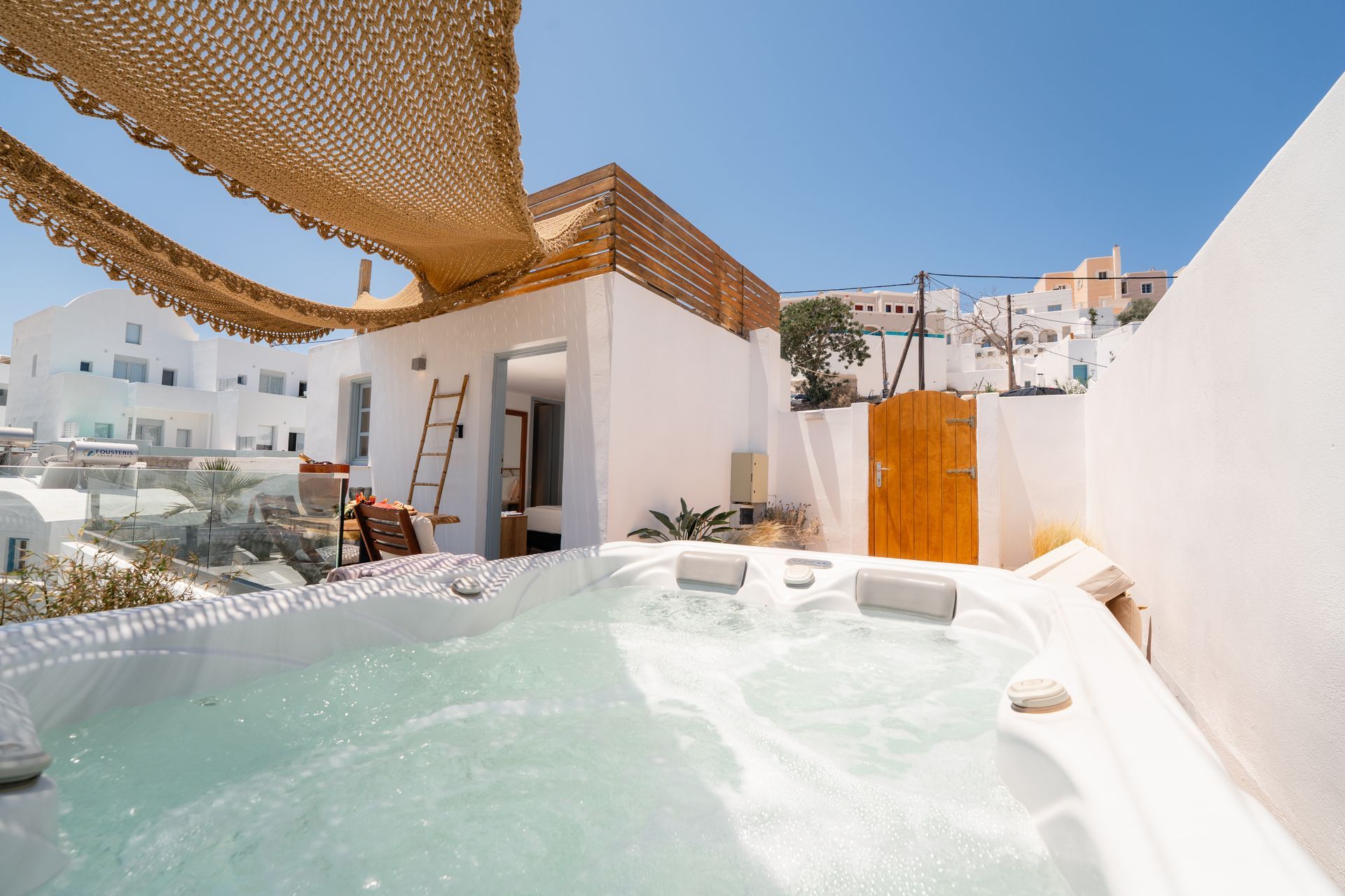 White-walled outdoor hot tub in Santorini, Greece, with partial shade cover and views of other buildings.