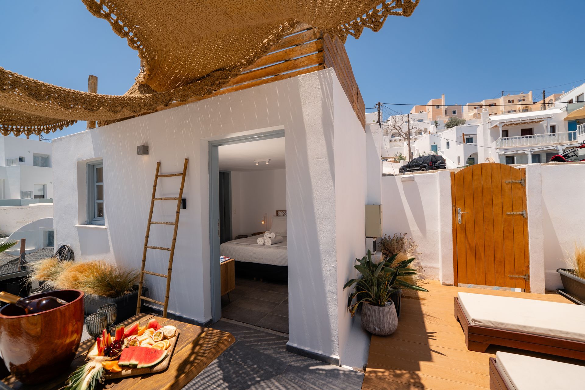 White building with open doorway to a bedroom, terrace with sunbeds, and wooden gate in a sunny Greek village.