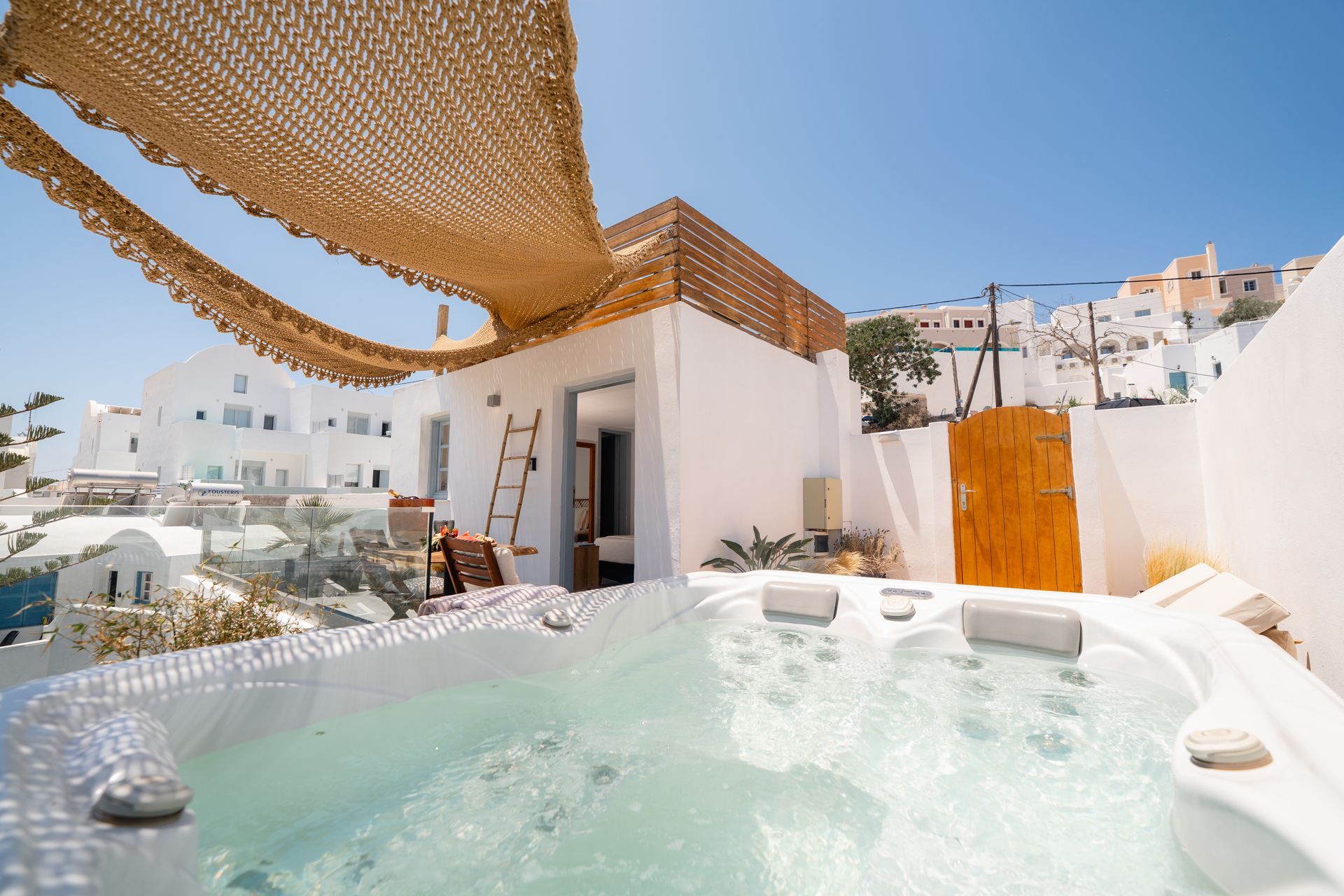 A private hot tub on a balcony in a Mediterranean village with white buildings and a woven shade canopy under clear sky.