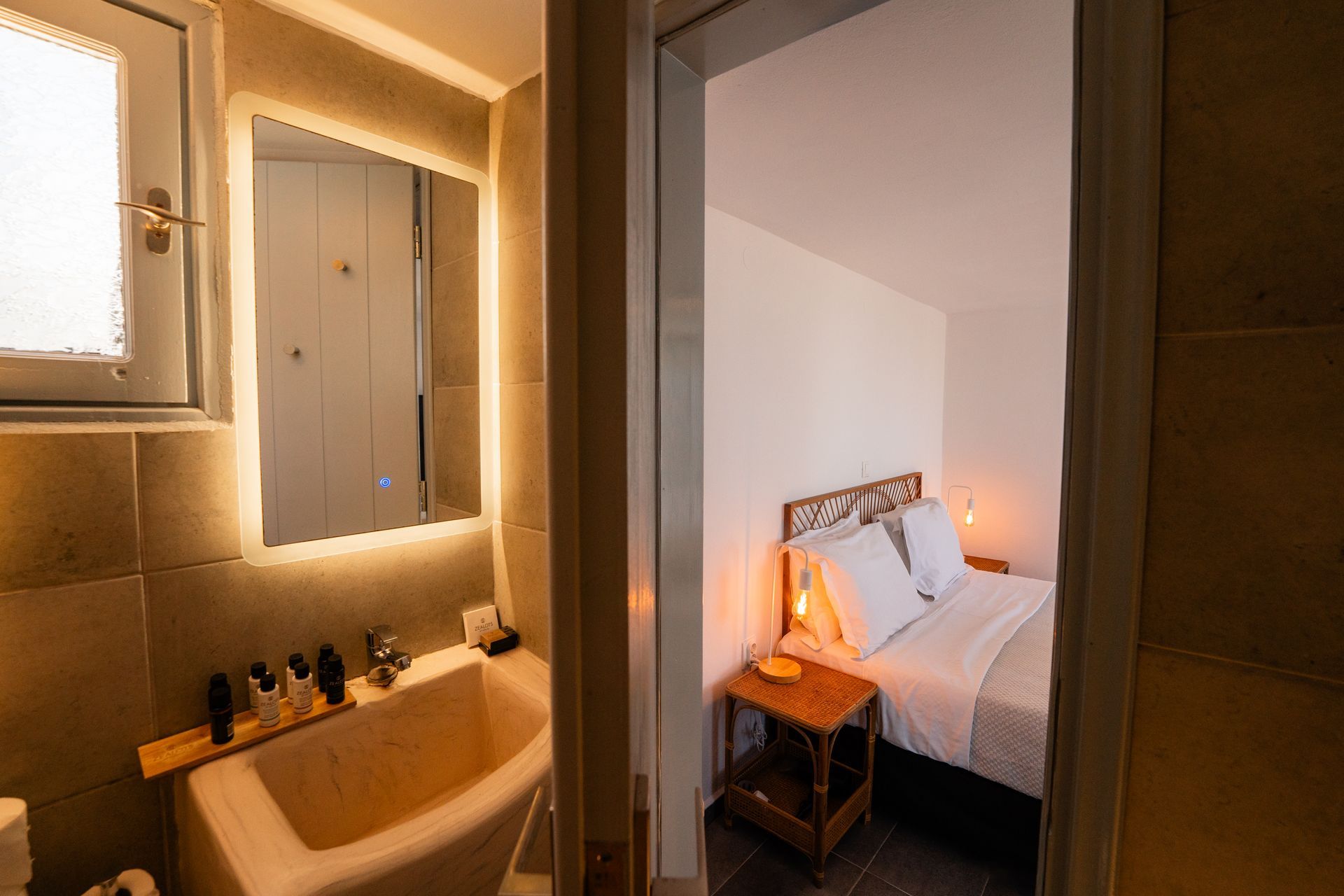 Bathroom with sink and mirror, doorway opens to bedroom with bed and nightstand.