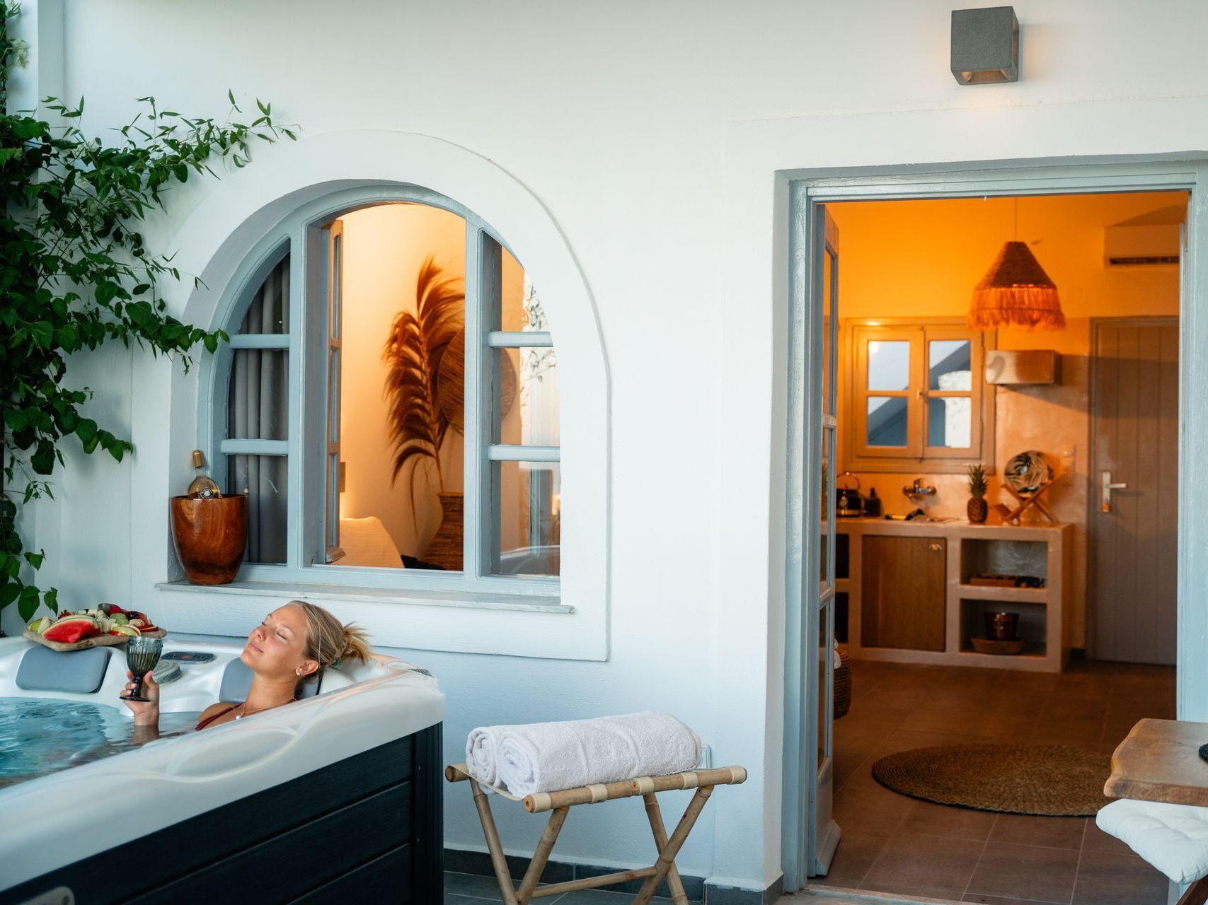 Woman relaxing in a hot tub outdoors, looking through a window into a warmly lit room.