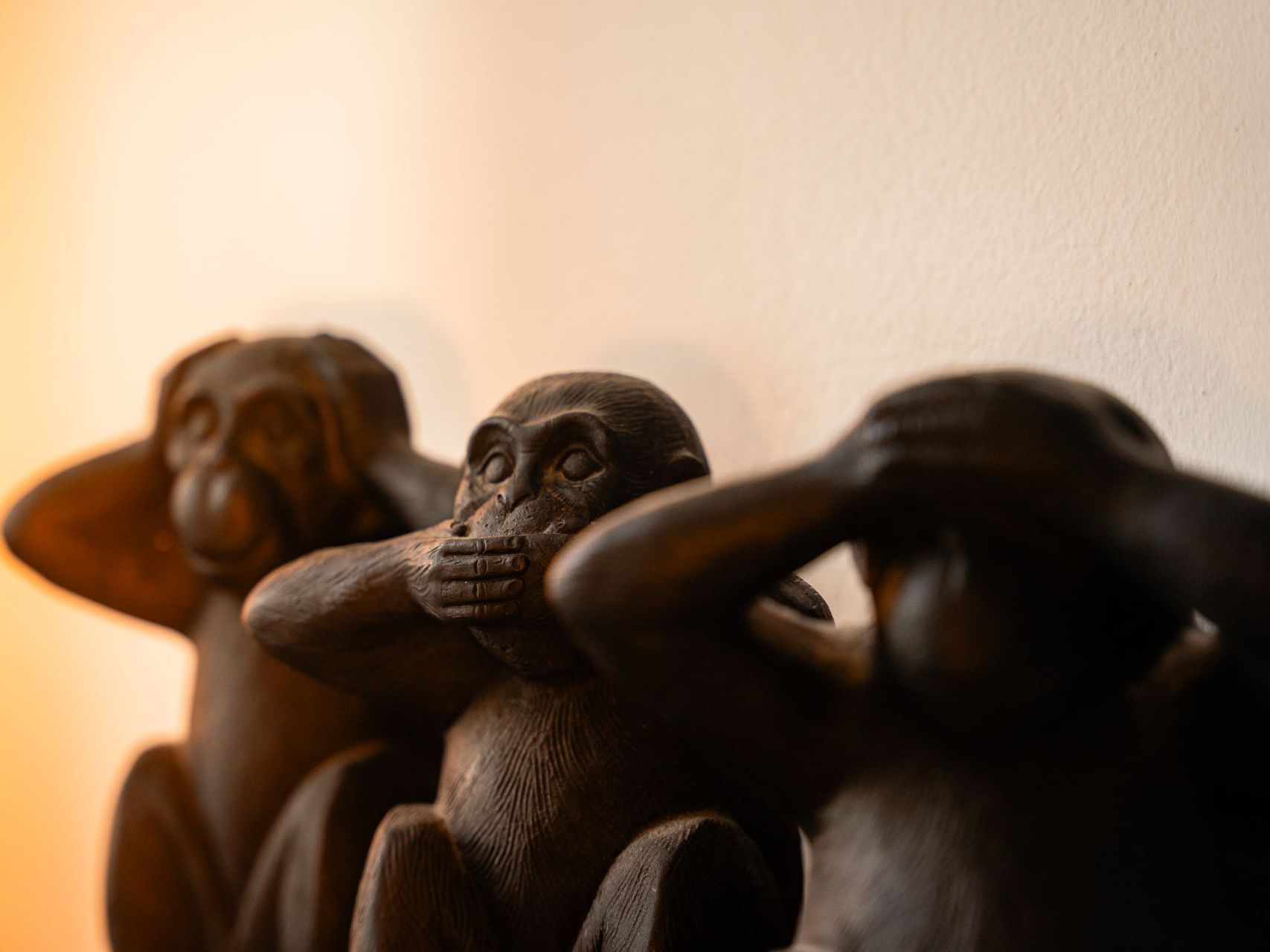 Three brown monkey statues: one covering ears, one mouth, one eyes; against a warm wall.