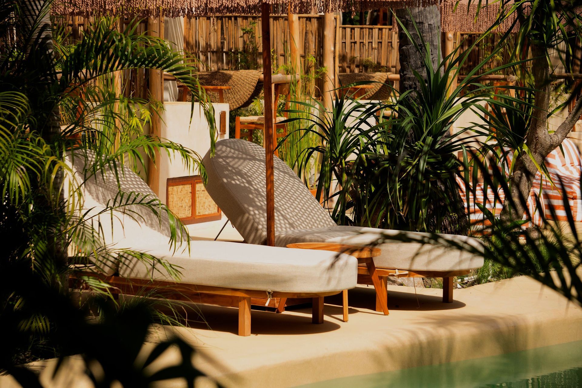 Two lounge chairs next to a pool, shaded by a straw umbrella, surrounded by tropical plants.