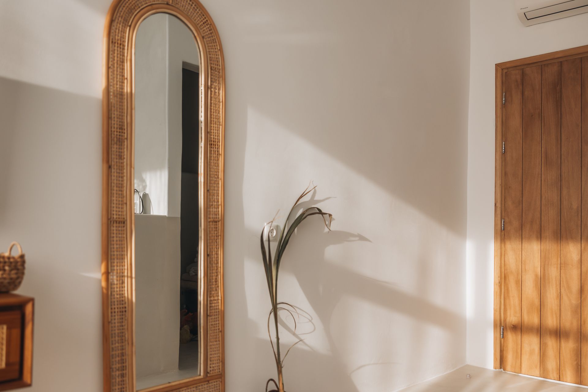 Tall, arched mirror with woven frame; white walls, wooden door, plant. Sunlight.