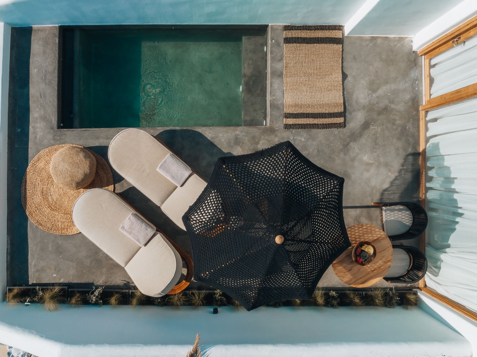 Overhead view of a pool area with two lounge chairs, umbrella, two straw hats, and small tables in Santorini.