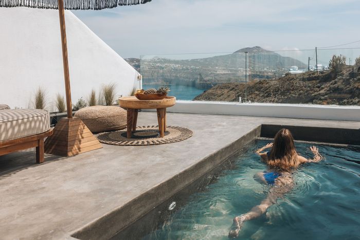 Woman in a pool overlooking the sea, next to a lounge area with a table and cushions.