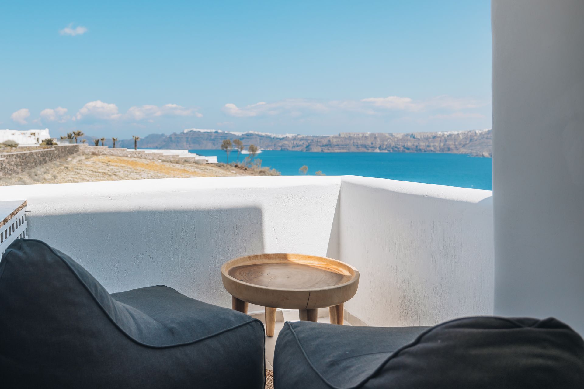 Balcony overlooking sea in Santorini; white walls, blue water, two beanbag chairs, wooden table.
