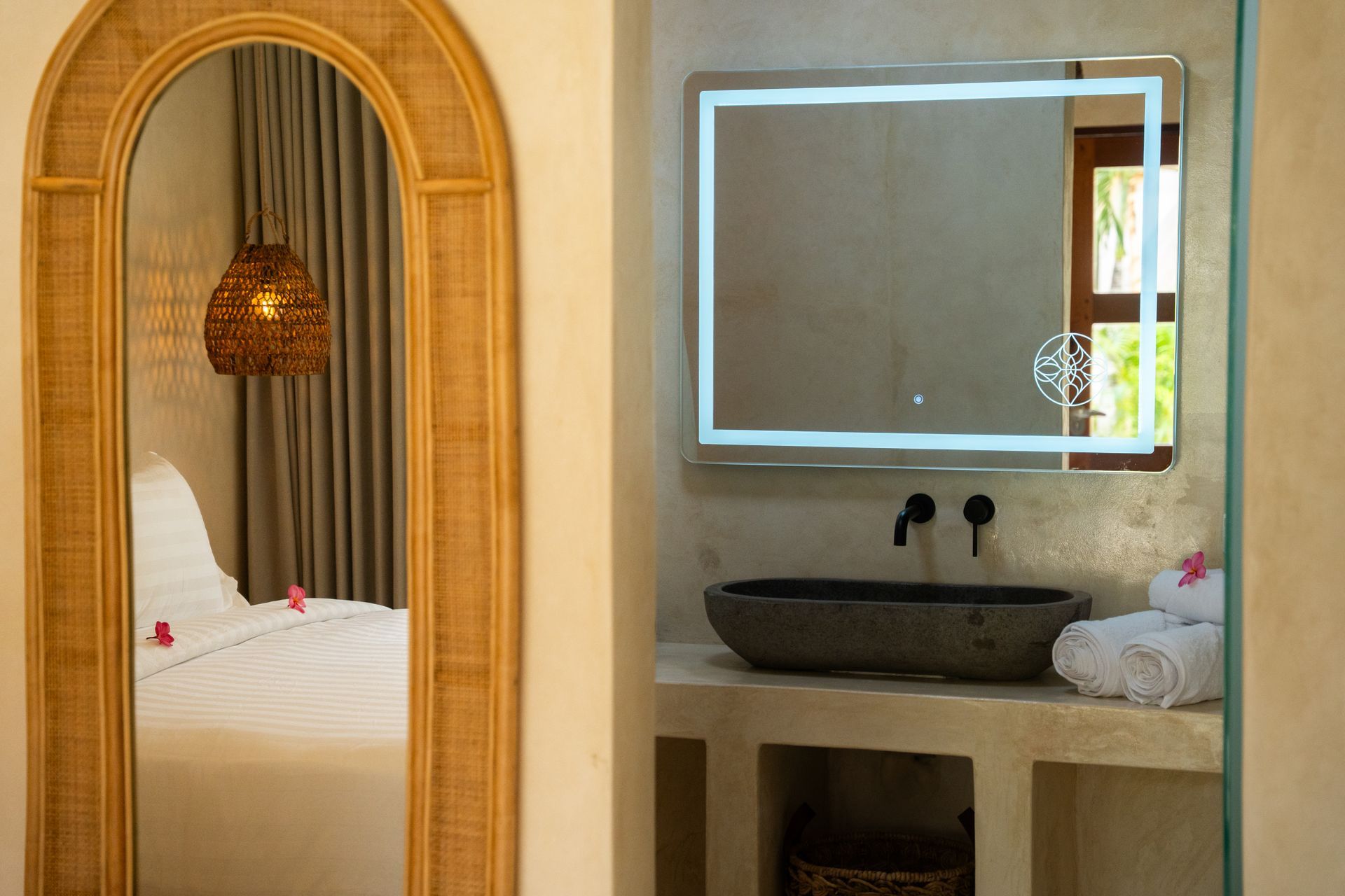 Bedroom with a rattan mirror, bed, and bathroom area with a lighted mirror and stone sink.