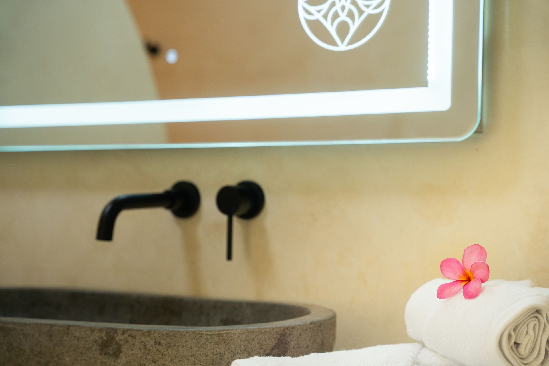 Bathroom sink with black faucet, large lit mirror, and rolled white towels with a pink flower.