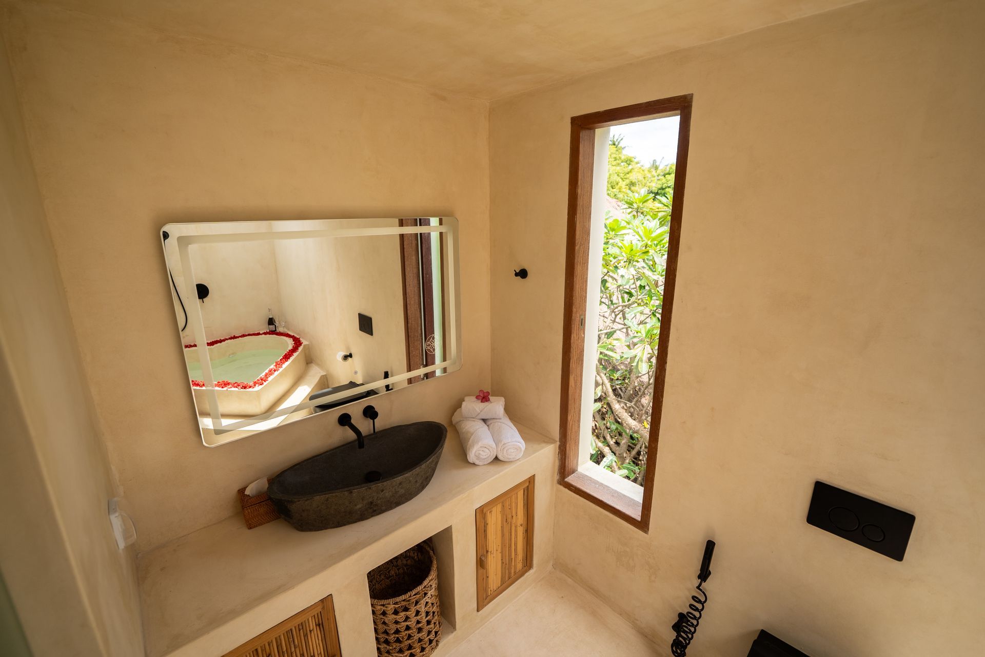 Bathroom with stone sink, mirror, small window, and a tub with rose petals.