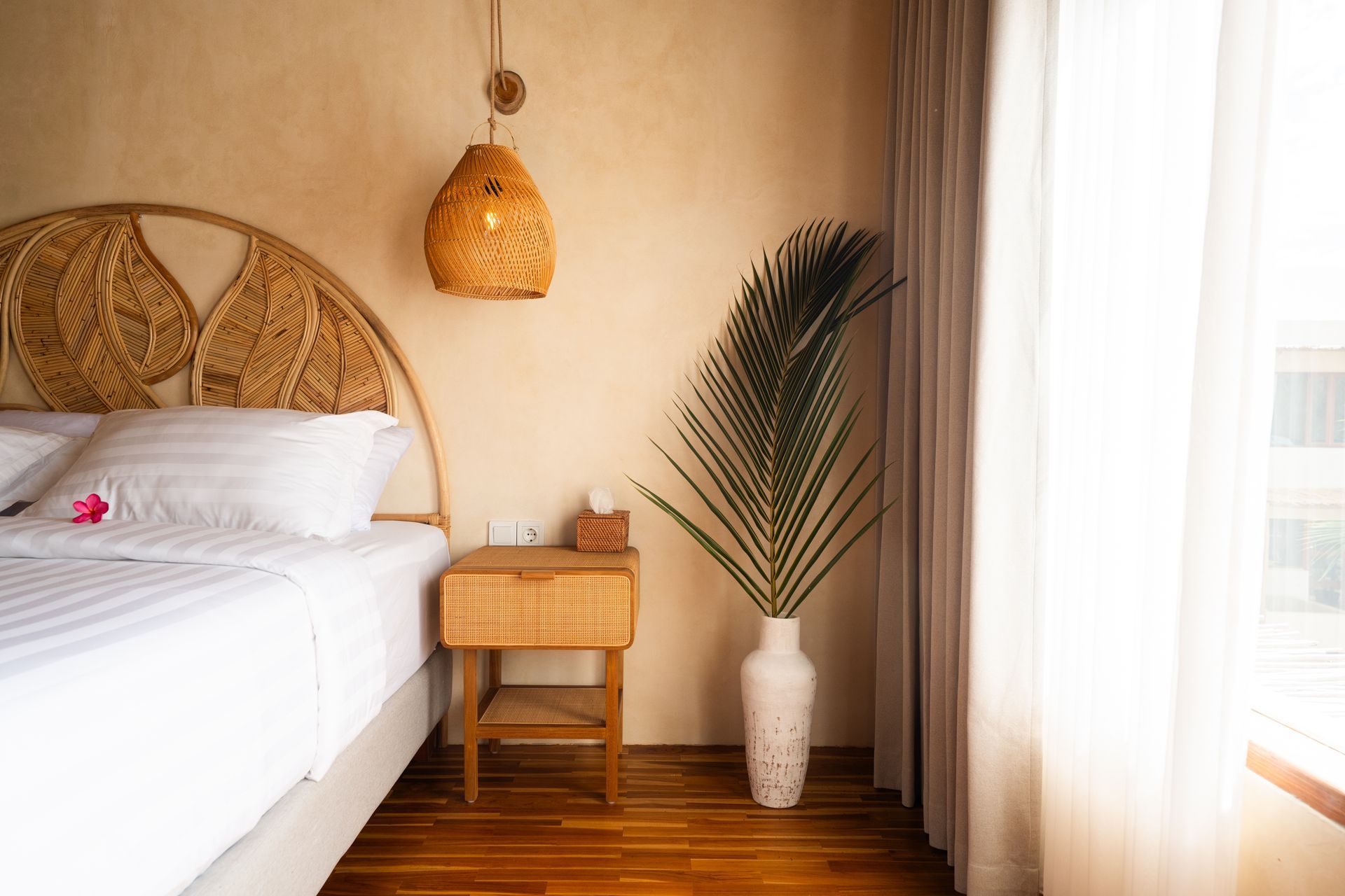 Bedroom with rattan headboard, bedside table, hanging light, vase with palm fronds, and sheer curtains.