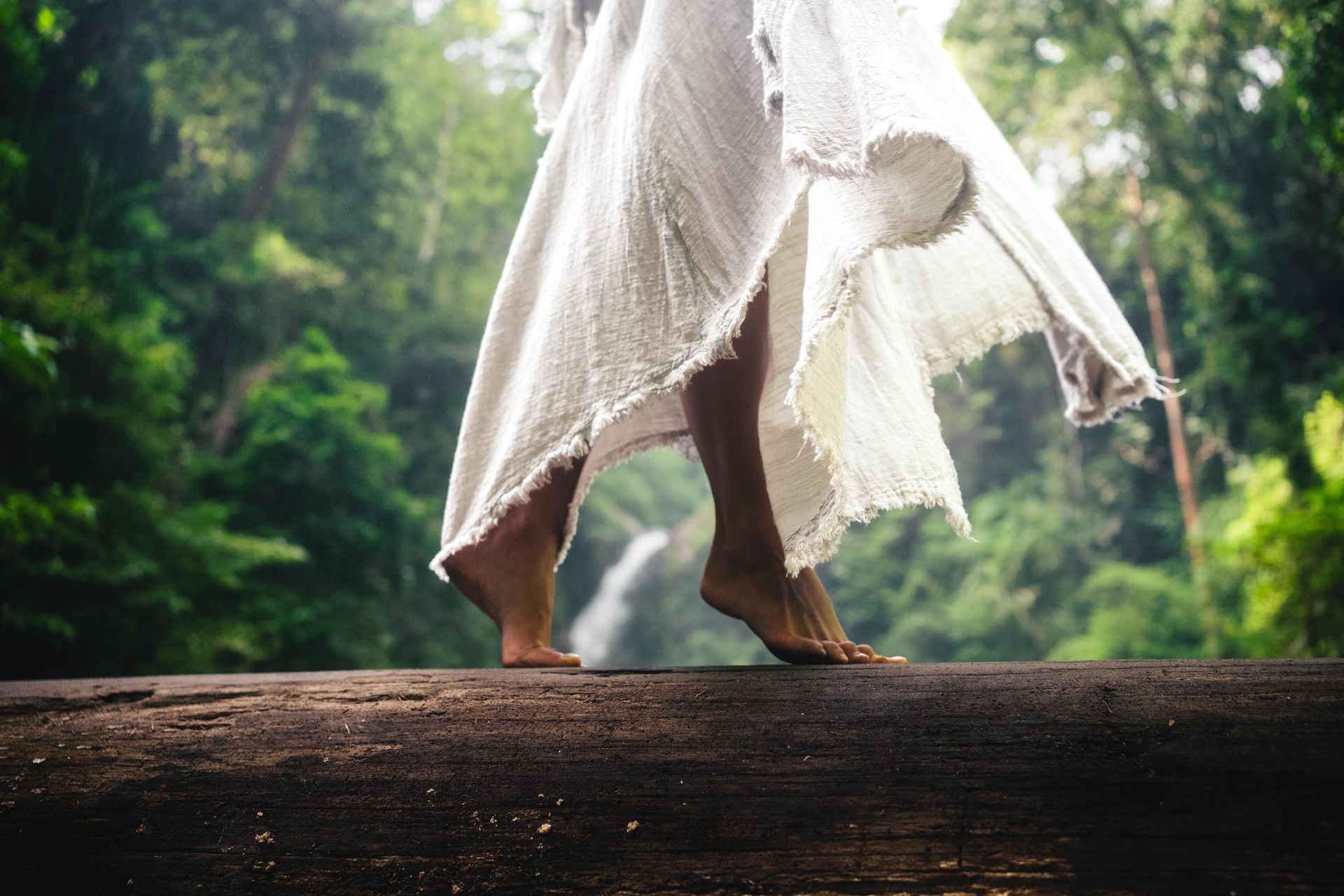 Bare feet walking on a log in a forest, wearing a white dress, with a waterfall in the distance.