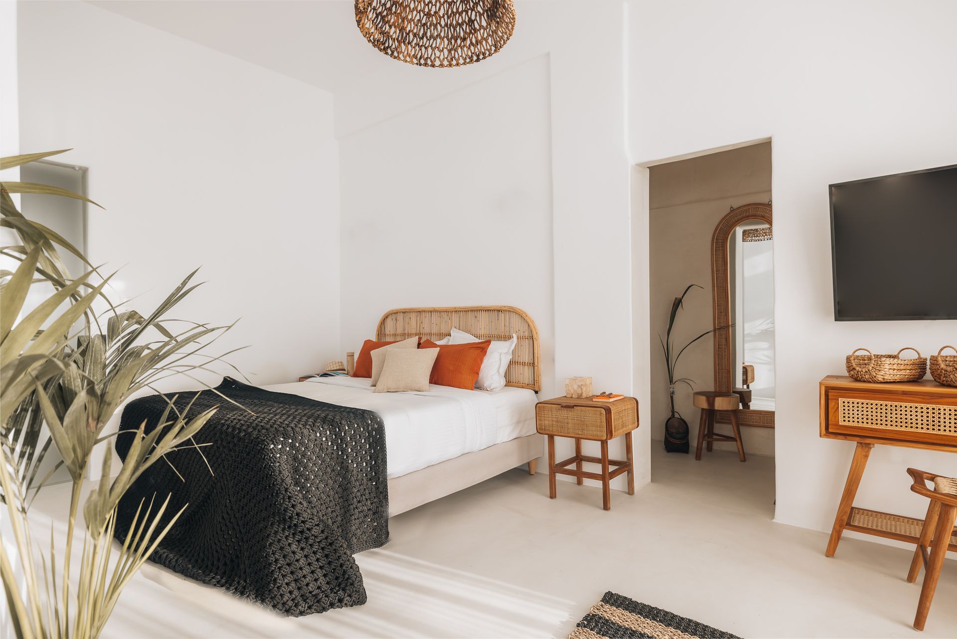 Bright bedroom with bed, woven headboard, side table, TV, and plants. White walls and floor.