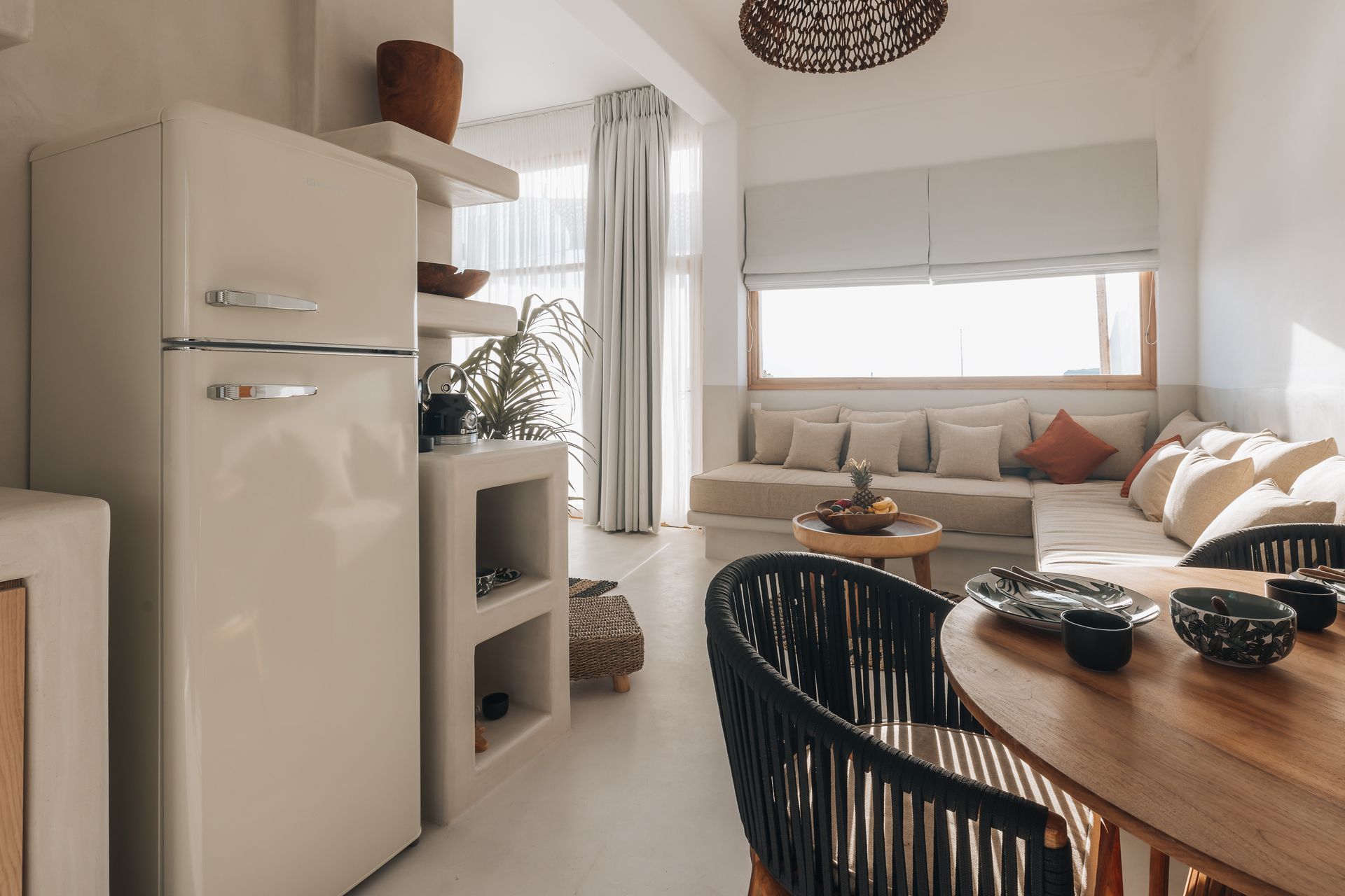 Cream-colored kitchen with retro fridge, built-in shelves, wicker chairs, and a tan sofa next to a window.