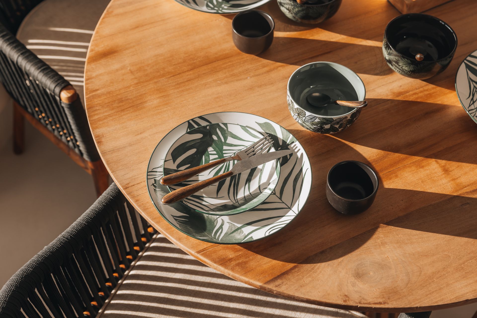 Wooden table with a plate, bowls, and cutlery, set for a meal. Sunlight and shadows across the table.