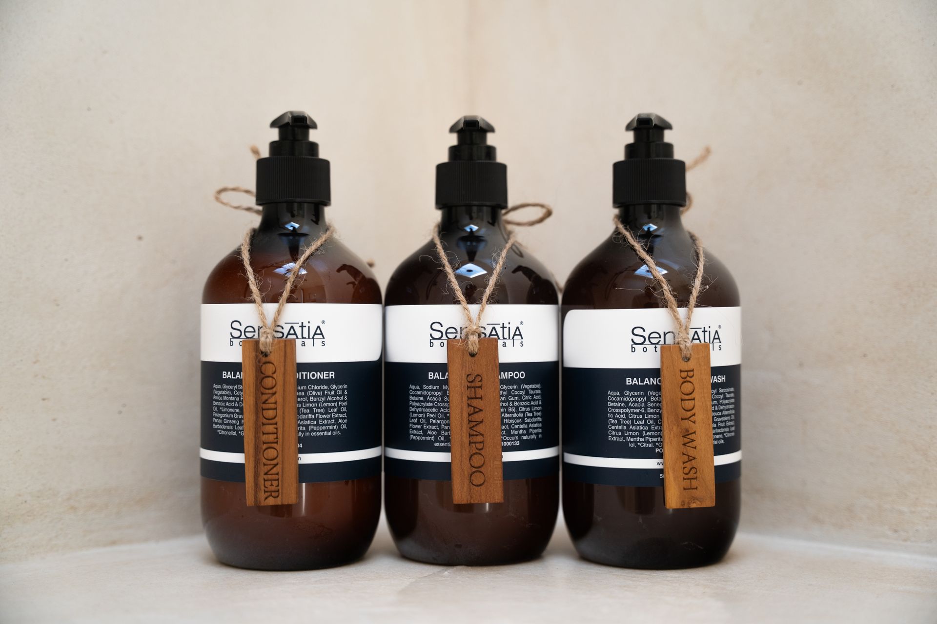Three brown shampoo bottles with black pumps, each with a wooden tag, set in a white corner.