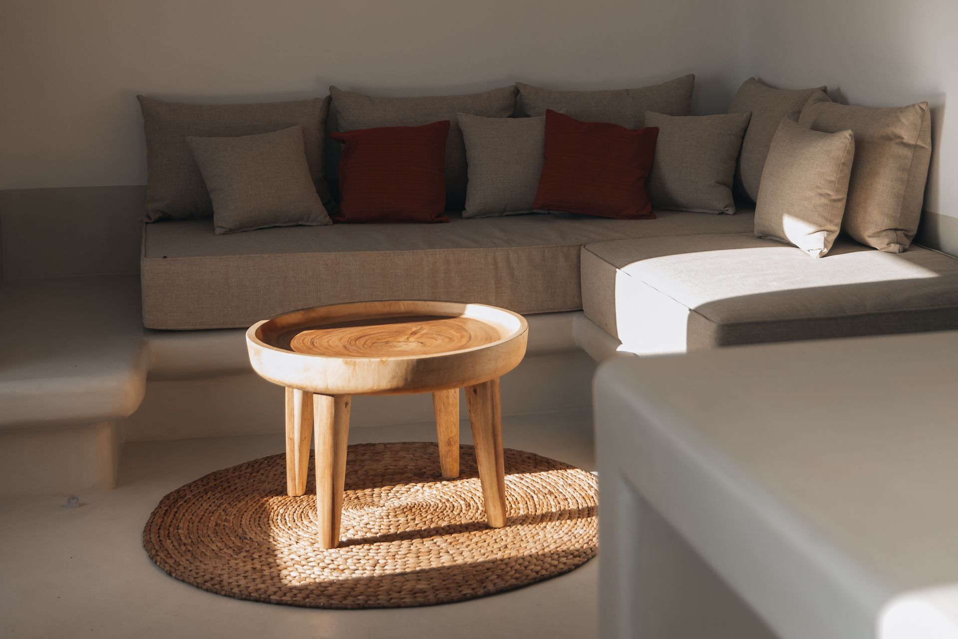A cozy corner with a beige sofa, red pillows, and a wooden table on a rug. Bright sunlight.