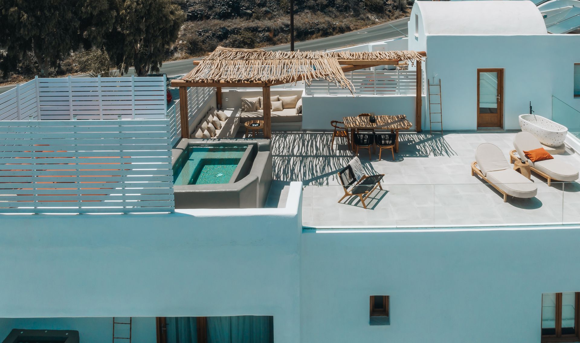 Rooftop with hot tub, seating, and sunbeds; white buildings in Greece. Sunny day.